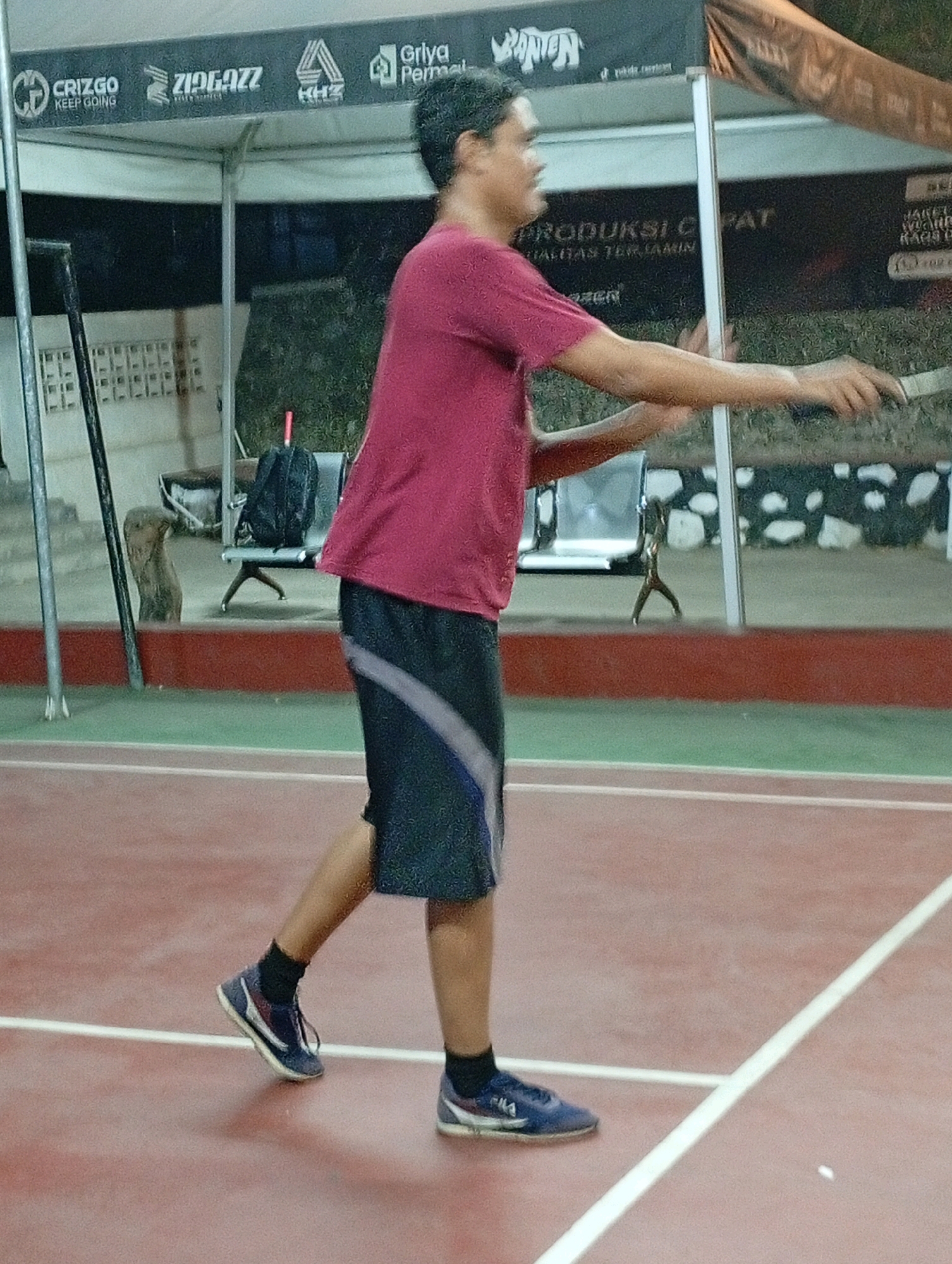 The image depicts a young male, appearing to be in his late teens or early twenties, playing badminton on an outdoor court. He is wearing a maroon t-shirt and dark athletic shorts with a light gray stripe down the side. His feet are clad in navy blue sneakers with white accents and black socks. He is in motion, holding a badminton racket, suggesting he is in the process of hitting or has just hit a shuttlecock. The court surface is reddish-brown with white lines marking the boundaries. In the background, there is a white tent structure with various logos, including "CRIZGO KEEP GOING," "ZIGAZZ," and "Griya Permat." Adjacent to the tent, a dark backpack rests on a metal bench. The overall environment appears to be a sports facility in Caringin, Indonesia, likely a public badminton court or club.