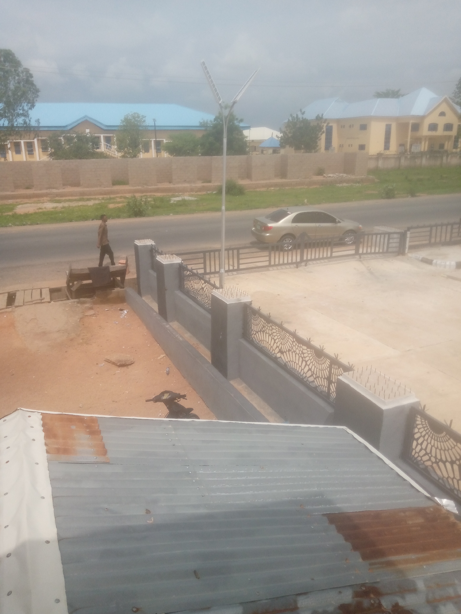 An outdoor, high-angle, slightly hazy, daytime shot shows a street scene in Jalingo, Nigeria. The foreground is dominated by a corrugated metal roof. Behind the roof, a gray concrete wall topped with black spike-like deterrents borders a paved area. A dark-colored motorbike is parked in the paved area. Beyond the wall and paved area is a road with a light-colored sedan driving on it. A man is walking on a dirt path beside the road. In the background, there are buildings with light blue roofs and yellow walls, separated by a low brick wall. A tall lamppost with solar panels stands near the road.
