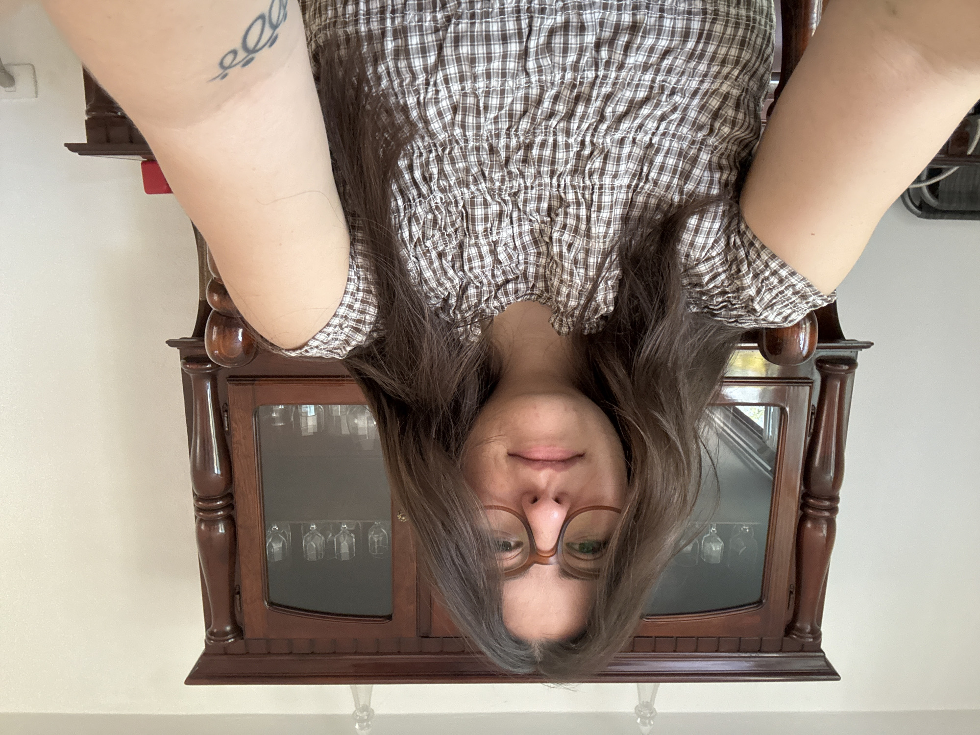 The image depicts a woman taking an overhead selfie, resulting in an upside-down perspective. When rotated correctly, the woman is seen from a slightly high angle, looking directly at the camera with a gentle, closed-lip smile. She has long, dark brown hair that frames her face and glasses with brown frames. She is wearing a brown and white checkered top, featuring a shirred or smocked bodice and elasticized sleeves.
On her visible left arm, a stylized script tattoo is visible on the inner bicep area.
Behind her, a dark wooden display cabinet with glass doors is visible. The cabinet has ornate, turned pillars on its sides and appears to be holding several stemmed glasses, hanging upside down, inside its upper section. The background also features a plain, light-colored wall. Bright, even lighting suggests it is daytime. The setting appears to be an indoor space, likely a home, within the context of Puebla, Mexico.