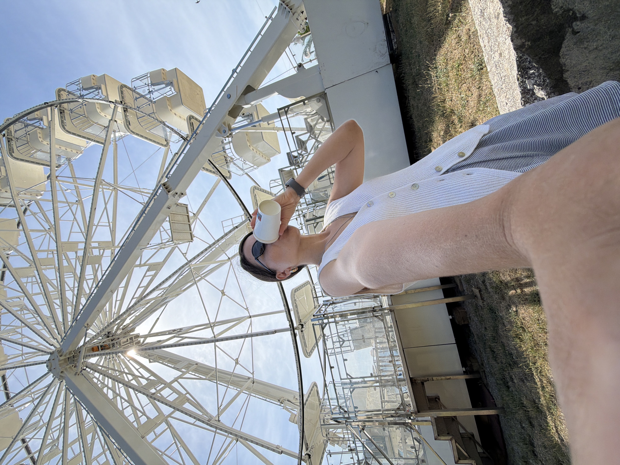 The image shows a person holding a cup and standing near a white Ferris wheel. The wheel is large and intricate, with enclosed gondolas. The person is wearing sunglasses, a light-colored top, and what appears to be a striped garment. The sky is blue with some visible light. There is grass and some structures near the base of the Ferris wheel. Based on the Ferris wheel's design and the surrounding structures, the location is likely in Budva, Montenegro.