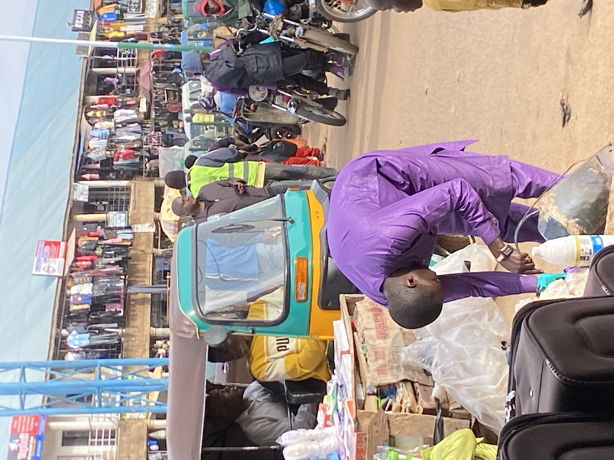 This media file depicts a busy street scene, likely a market, in Bukuru, Nigeria. The image is vertically oriented and shows several individuals and various objects, suggesting a bustling commercial environment.
In the foreground, a person wearing a vibrant purple robe is bending over, appearing to interact with something on the ground. Their posture suggests focus or engagement in a task. Adjacent to them, there are several pieces of luggage, including dark suitcases, indicating travel or the movement of goods. A three-wheeled vehicle, commonly known as an auto-rickshaw or "keke," is partially visible, its teal and yellow colors adding a splash of contrast to the scene.
The background reveals a dense collection of market stalls, overflowing with merchandise. Items such as clothing, bags, and other assorted goods are displayed, creating a sense of abundance and activity. Several people are visible within the market, some dressed in casual attire, others in work-related vests like the neon green one worn by a person in the mid-ground. Motorcycles are also present, suggesting transportation and the movement of people and goods within the market.
The lighting suggests it might be daytime, likely under clear or partly cloudy skies, casting distinct shadows. The overall atmosphere appears lively and energetic, typical of a busy marketplace. The presence of diverse items and the sheer number of people engaged in various activities point to a functioning commercial hub. The text "Topbell" is visible on a sign in the lower left portion of the image, possibly indicating a business or brand. Another sign with "WORLD" is also partially visible.