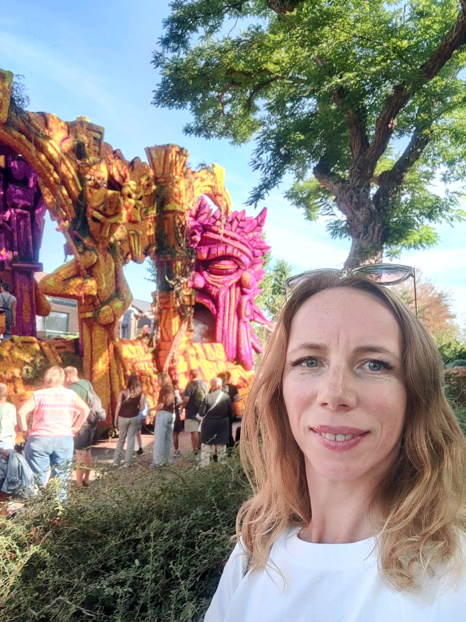 The image is a selfie taken outdoors in Zundert, Netherlands, during the daytime. In the foreground, a woman with blonde hair and blue eyes is smiling at the camera. She is wearing a white t-shirt and sunglasses are perched on her head. Behind her, a large, colorful float decorated with a skull motif and adorned with pink and orange flowers is visible. The float appears to be part of a parade or festival, as other people are seen in the background observing it. The float has intricate carvings and is made of many individual components. The environment suggests a sunny day with a clear blue sky and green trees in the background.
