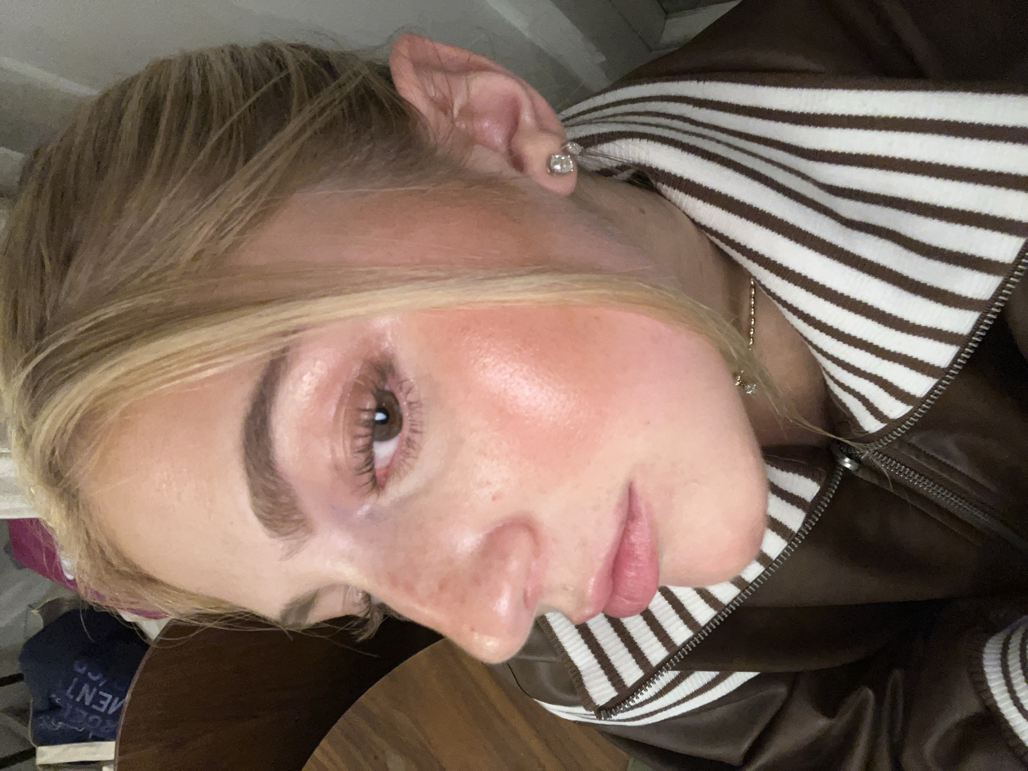 A close-up, portrait-style image shows a woman with light blonde hair, fair skin, and brown eyes. She is wearing a brown jacket with white stripes on the collar and what appears to be a chain necklace. She has a stud earring in her left ear. The background is out of focus. The location is not discernible and there is no indication of Paris, France.