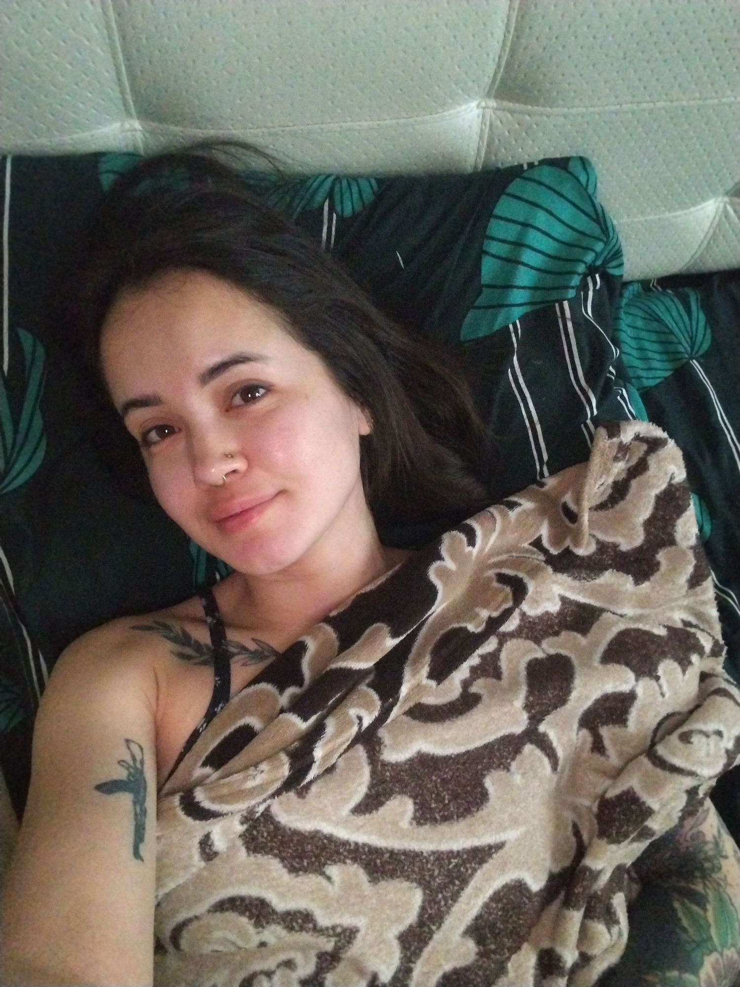 A woman lies in bed, covered partially by a brown patterned blanket. She has dark hair, a nose ring, and visible tattoos on her upper left arm and chest/shoulder area. The bed has a white headboard and is covered with a dark patterned bedsheet with green and white accents. The location is not provided in the image.