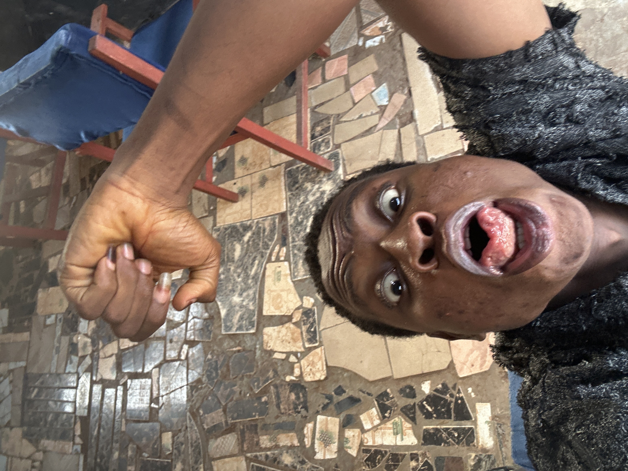 A person is holding the camera above their head, pointing downward at their face and body. The person's mouth is open with their tongue visible. They are wearing a gray shirt. Their right hand is held up, with fingers curled into a fist. A blue cushioned chair with a red frame is in the upper-left corner. The floor is made of broken tiles. Location: Unknown, Unknown.