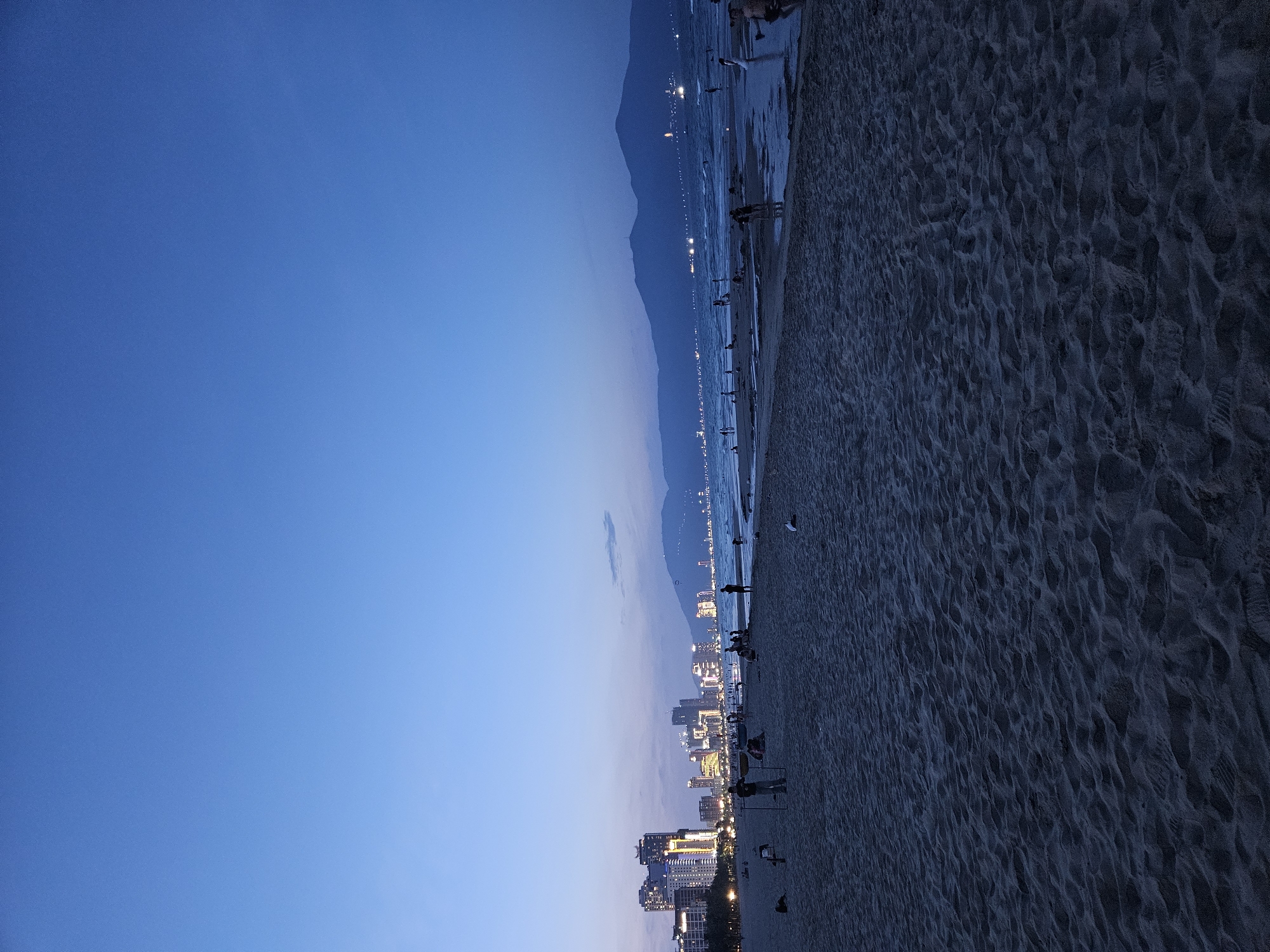 This is a low-angle, medium shot of a beach in Da Nang, Vietnam, at dusk. The sand in the foreground is dark and textured with footprints. In the midground, several people are scattered across the beach and wading in the shallow water. Some are silhouetted against the sea. Beyond the beach, the city skyline is visible, with illuminated high-rise buildings. In the background, mountains loom under a deep blue sky, with a faint haze of clouds. The sea is calm and reflects the city lights.