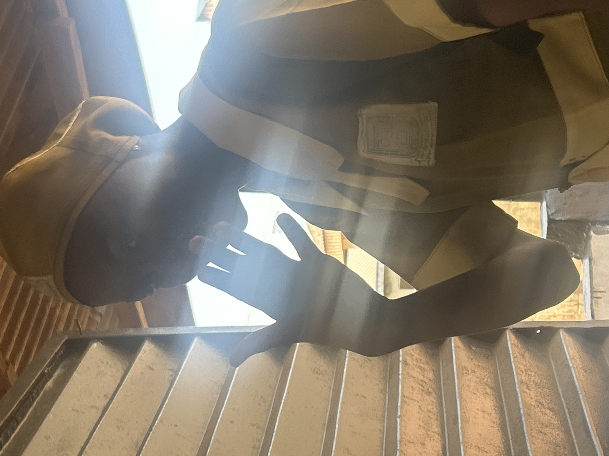A person wearing a tan cap and a brown shirt with white trim is looking down at a gray metal grate. Their hand extends over the grate, partially obscuring the view. The grate appears to be situated on the floor or ground level. The location is Unknown, Unknown.