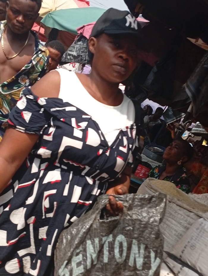The image depicts a bustling outdoor market scene in Onitsha, Nigeria. In the foreground, a woman faces slightly towards the viewer's right, making eye contact with the camera. She wears a white sleeveless top underneath a dark blue, white, and red patterned off-shoulder dress. A black baseball cap with a white "NY" logo covers her head. She holds a crumpled grey plastic bag displaying the white text "GENTONY" on its surface. To her left, slightly behind her, a shirtless man with a gold-colored chain around his neck wears an open patterned shirt and looks downwards. Another man is partially visible behind him. The background reveals a crowded market environment with multiple individuals, stalls, and various goods. Overhead, sections of green and pink fabric awnings provide shade. On the right side, sacks of what appear to be dry goods are stacked, and another woman in the far background looks upwards.