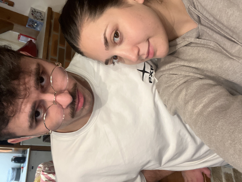 A man with round glasses and a mustache wears a white t-shirt with a small black logo. A woman rests her head on his shoulder, wearing a light gray hoodie. They both look towards the camera. A kitchen corner is visible in the background.