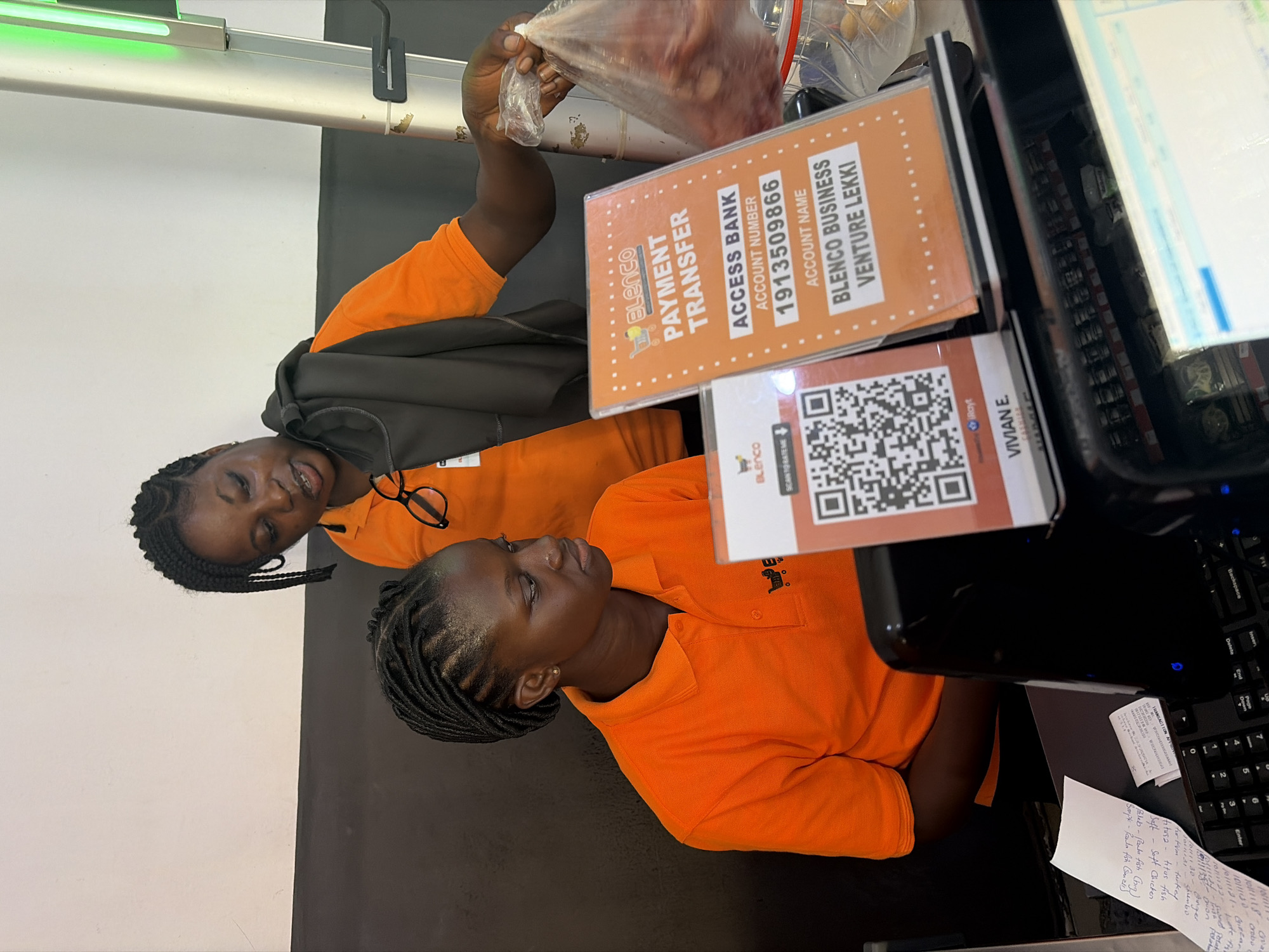 Here's a detailed description of the image:
**Scene:** A checkout counter at what appears to be a Blenco Business Venture Lekki store in Igboefon, Nigeria.
**People:** Two Black female employees, both wearing orange polo shirts, are present. One is handling a plastic bag of what appears to be a food product.
**Objects:**
* Cash register and keyboard
* Signs indicating payment transfer information for Blenco:
* Access Bank account number: 1913509866
* A QR code
* A receipt or order note with handwritten items, including "Soft Chicken," "Titus Fish," and "Kim Turkey."
* A pair of eyeglasses
**Actions/Interactions:** One employee is handing the plastic bag, presumably to a customer. The other employee is at the cash register.
**Environment:** The lighting is bright. The background is a combination of white and gray walls.