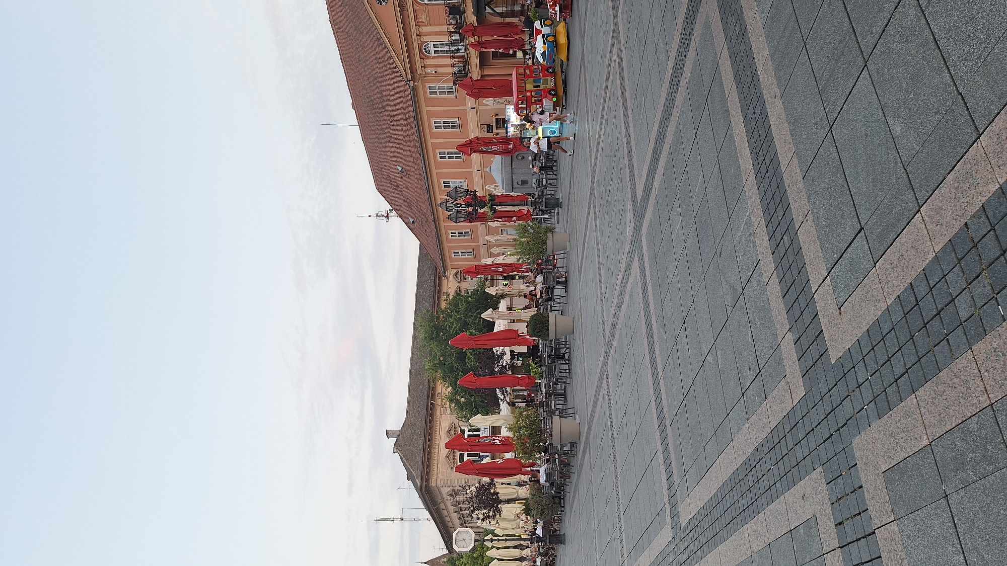 The image shows an outdoor cafe scene in Sombor, Serbia, specifically in the main city square, Trg Svetog Trojstva (Holy Trinity Square). The foreground is dominated by a paved courtyard with a geometric pattern of dark grey and reddish-brown tiles. In the mid-ground, tables and chairs are set up under numerous red umbrellas, suggesting a lively cafe environment. People are seated at some of the tables, engaged in conversations or enjoying their meals. To the left, a building with a prominent clock tower is visible, and further back, other buildings with traditional architecture form the backdrop of the square. To the right, a brightly colored children's amusement ride, resembling a train and a car, is present. The sky is a pale blue with light clouds, indicating it is daytime, possibly late afternoon or early evening given the soft lighting. The overall impression is that of a typical European city square that serves as a social gathering place.