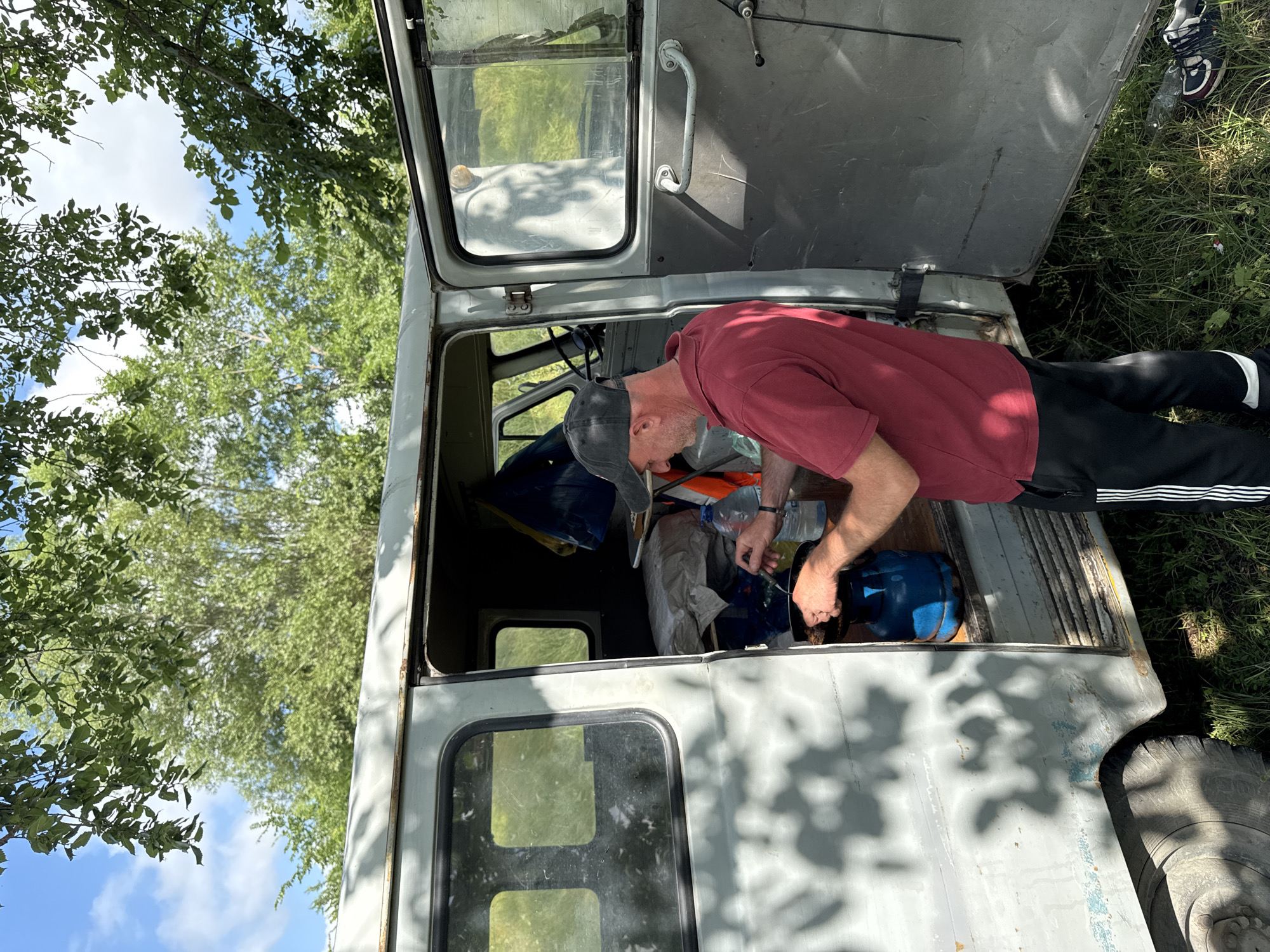 A middle-aged man wearing a maroon polo shirt and a black baseball cap is seen from the side, preparing food outdoors. He stands beside an open, light gray van with its rear door ajar. The man is actively engaged in cooking, holding a utensil over a small pan placed on a blue gas cylinder. A plastic bottle of water is also visible nearby. The scene appears to be set in a grassy area with trees and foliage in the background, suggesting an outdoor location. The light gray van has visible wear and tear, with some rust and peeling paint on its exterior. The overall environment indicates a casual, possibly camping or roadside setting. There is no information to confirm or deny the location being Khidiskuri, Georgia.