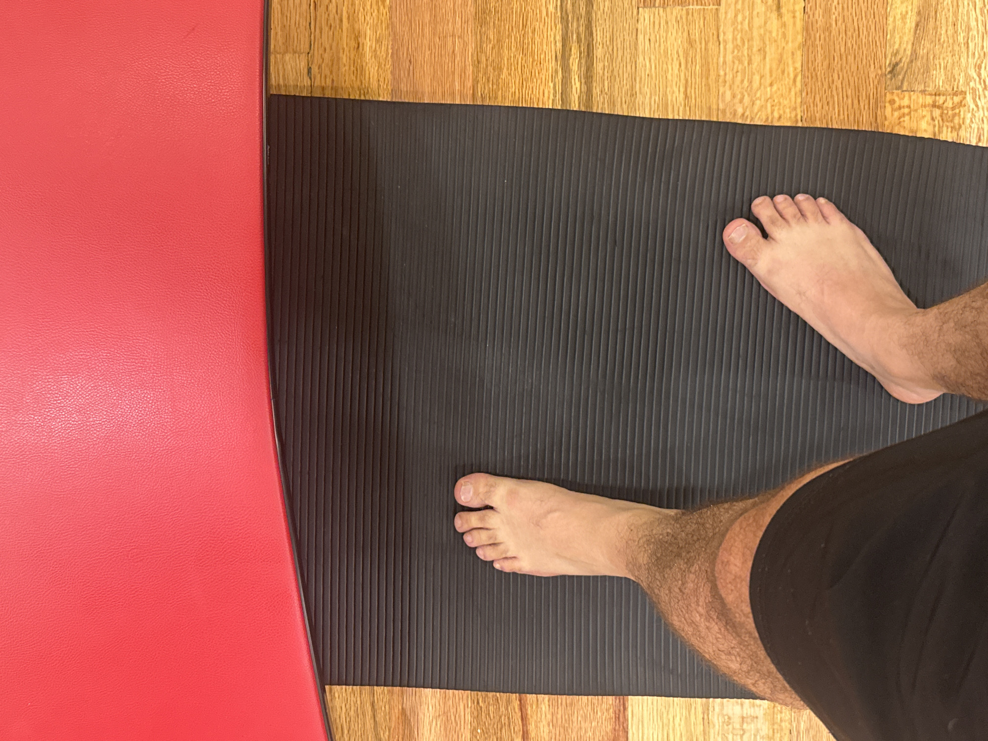 The scene depicts a person standing on a black yoga mat with bare feet. The mat is placed on a wooden floor and is partially adjacent to a red surface, possibly another mat or object. The person is wearing black shorts. The setting is indoors, with no further environmental cues to identify the location as Puebla, Mexico.