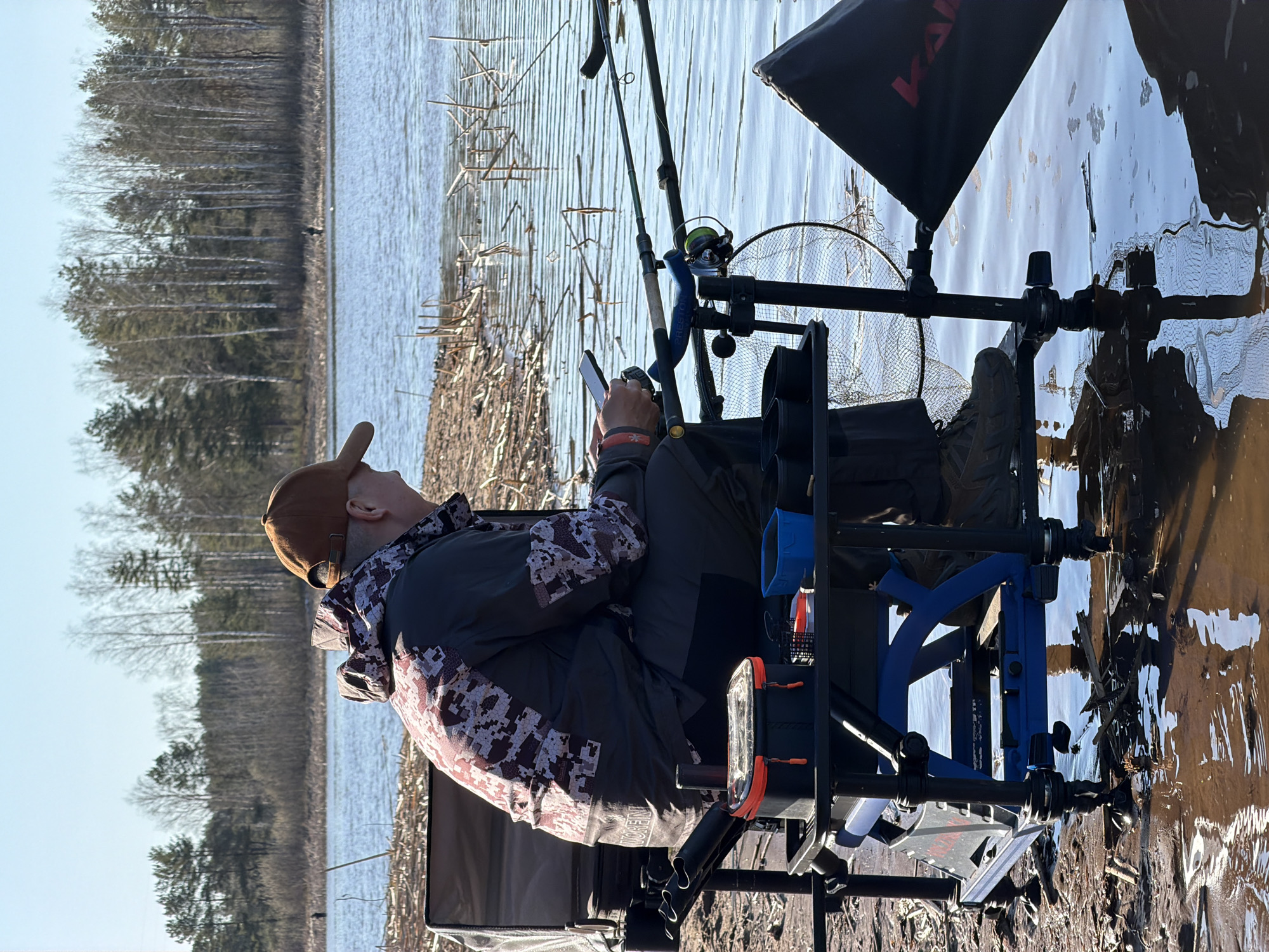A man is sitting in a fishing chair by a body of water. He is wearing a brown cap, a gray jacket with a camouflage pattern, and dark pants. He is holding a cell phone in his hands. Fishing rods are set up beside him. The chair is black and blue, and it has various attachments, including a black canopy with red lettering. A fishing net is positioned behind the chair. The water is rippled, reflecting the sky and the surrounding trees. Across the water, a line of trees can be seen. Reeds and branches are visible along the shoreline. The location is in the city of Chaikovsky, Russia.