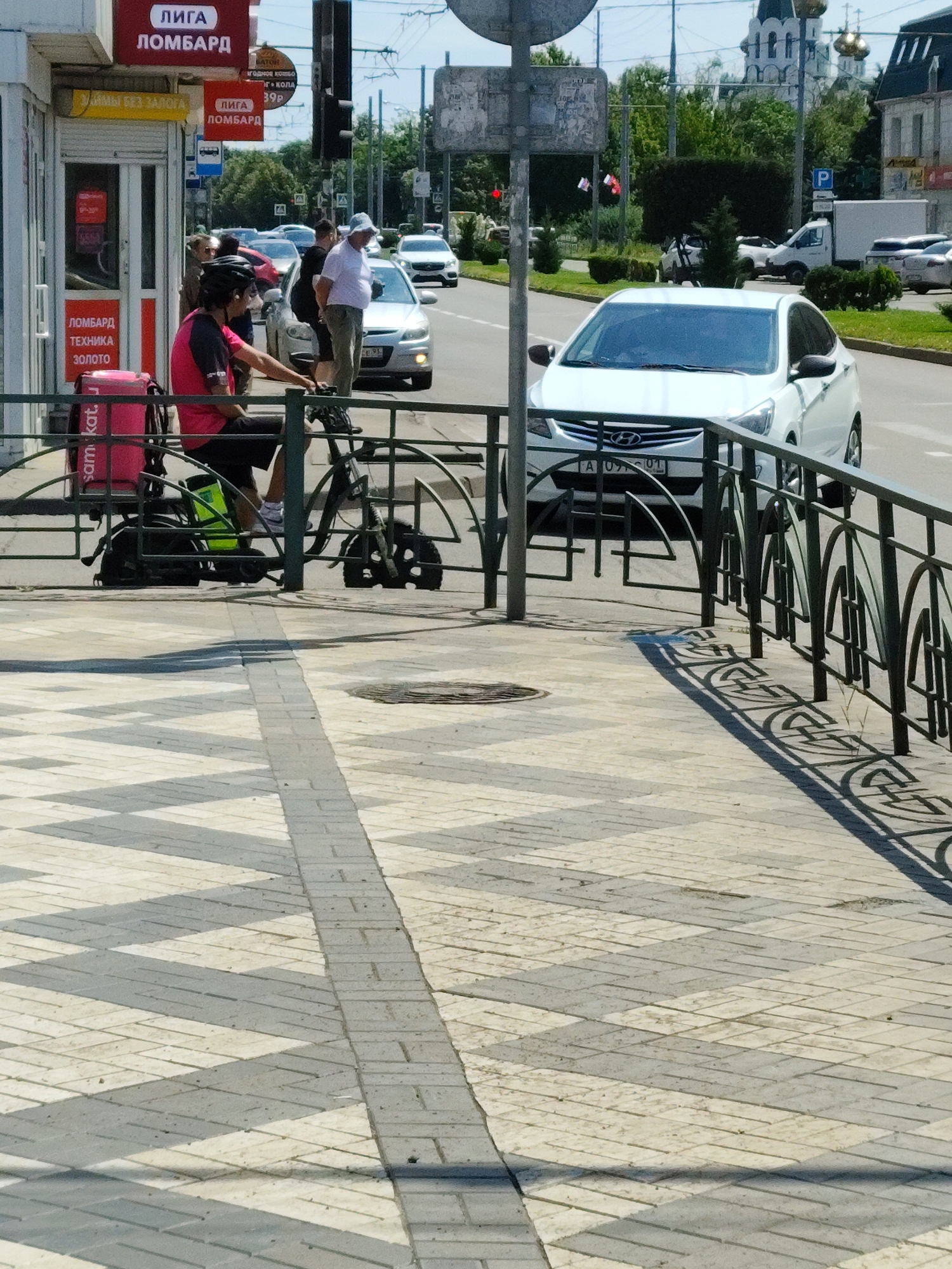 A courier on a scooter with a bright pink delivery bag is positioned behind a metal fence. The scooter has a green water bottle attached to its frame. To the left of the courier, a building with red signage is visible. The building has a small kiosk-like structure with a glass door and a red object inside. Above the kiosk, signs with Cyrillic text are displayed. In the background, a white car with a license plate starting with "A U9" is parked on a street. Other vehicles and trees line the street, and a church with golden domes is visible in the distance. The pavement in the foreground features a pattern of light and dark grey interlocking bricks, creating a zigzag design. The scene appears to be a sunny day in an urban environment.
