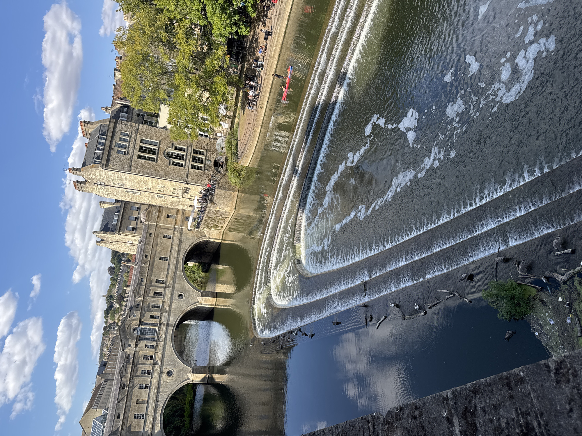 This is an eye-level shot of a cascading waterfall on the River Avon in Bath, United Kingdom. The waterfall, made of concrete steps, is in the foreground and fills the right half of the image. The left side of the image is dominated by the reflection of the sky and the Pulteney Bridge, a historic stone bridge with shops built into its structure, spanning the river. The bridge is in the middle ground, with its arched structure clearly visible. The water is a murky green color, and there are several pieces of driftwood and trash visible in the lower left corner. A few ducks are scattered in the river. In the background, on the right bank, are several buildings with traditional stone facades and pitched roofs, with lush green trees bordering the river. A few people are visible on the riverbank, some seated at tables outside a building, and one person is on a paddleboard in the river. The sky is blue with fluffy white clouds.