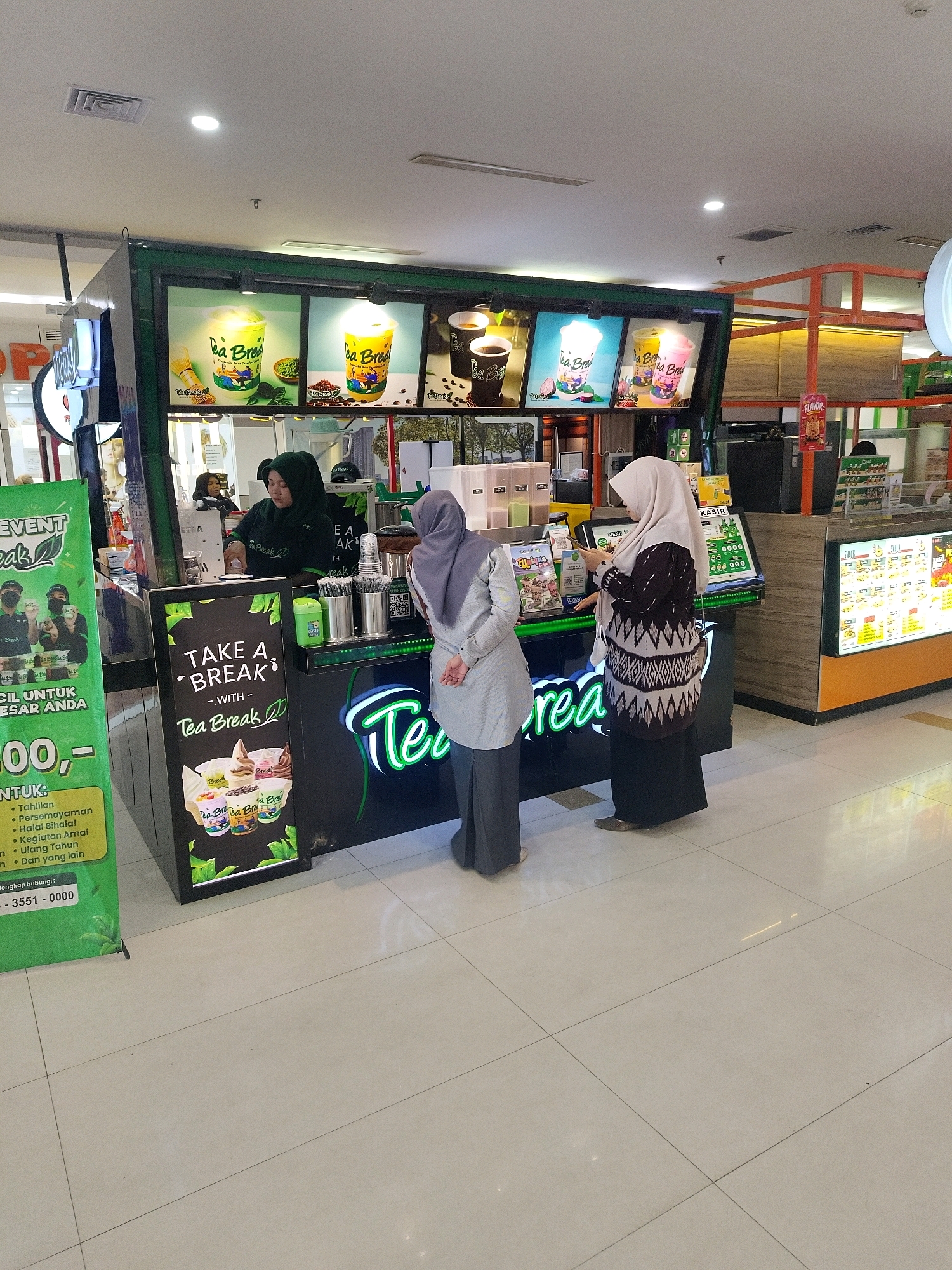 The image depicts an outdoor food stall in what appears to be a mall or a busy shopping area in Mojokerto, Indonesia. The stall, branded as "Tea Break," is set up with a green and black aesthetic. It features illuminated menu boards displaying images of various beverages, including iced teas and coffee drinks.
Two female employees are visible behind the counter, one wearing a dark green hijab and uniform, actively serving customers. The other, wearing a light grey hijab and a patterned dress, is engaged in conversation with a customer. Two customers are present; one is wearing a grey hijab and a dark patterned outfit, looking at a tablet or phone. The other customer is partially obscured but appears to be in the process of ordering.
In the foreground, a large green banner with text and images related to "Tea Break" is displayed, highlighting promotions and contact information. The floor is tiled in a light color, reflecting the overhead lighting. The overall impression is that of a bustling commercial environment where a beverage vendor is catering to shoppers.