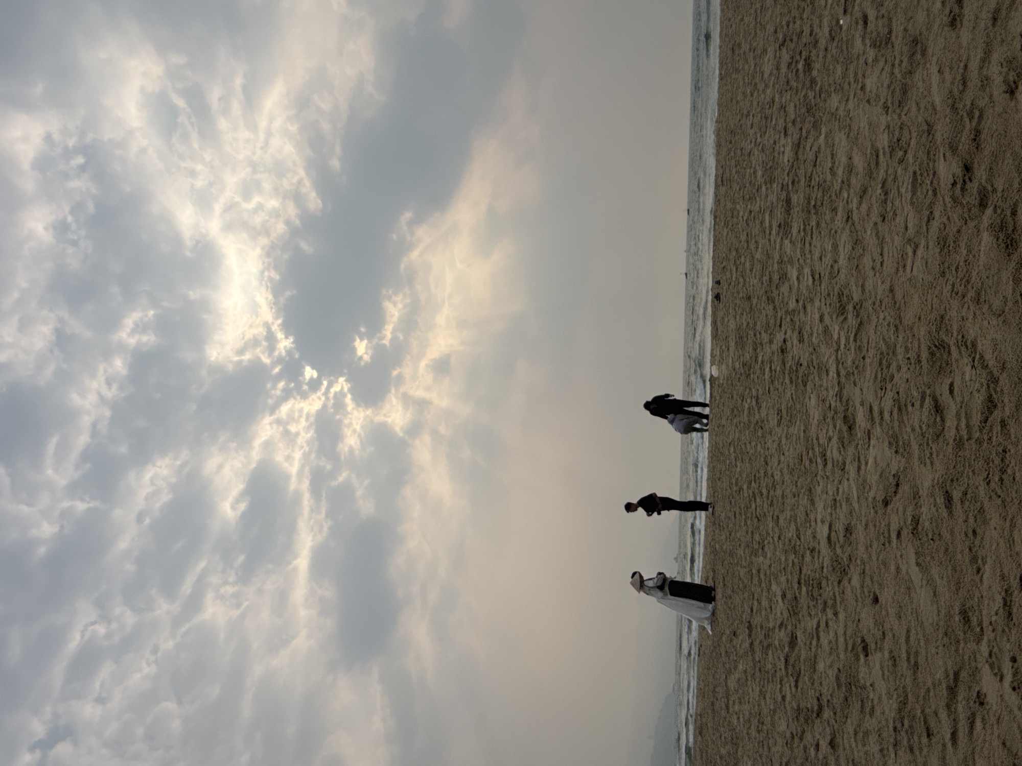 The image shows a beach in Đà Nẵng, Vietnam. Three people are on the beach near the water's edge, with the sea to their left and the sand stretching out to the right. The sky is overcast with a mix of grey and bright clouds. One person is wearing what appears to be a white dress or robe with a head covering. Another is wearing dark clothing. The third person seems to be crouched down.