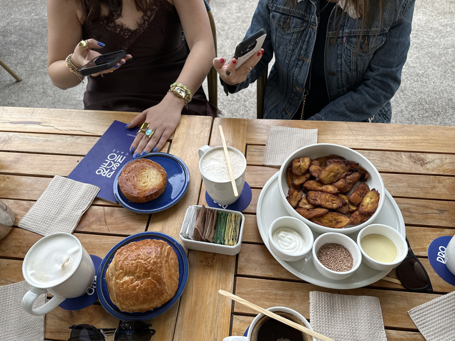 An overhead view captures a wooden slatted table at Preco Café in Mexico City, Mexico, set with food and beverages for two individuals.
The table holds a large croissant and a smaller laminated pastry, each on a blue ceramic plate. A prominent white oval platter presents a central bowl filled with fried plantains, flanked by three smaller white bowls containing white cream, brown sprinkles, and a light yellow sauce. Beverages include two white mugs with frothy drinks and one dark mug with black coffee, each with a wooden stirrer. A white container holds assorted sweetener packets. A blue folded menu partially displays "PRECO RESO CAFE."
Two individuals are partially visible on the right. The upper individual, wearing a dark brown lace-trimmed top, has hands with blue nail polish, multiple rings (one featuring a large turquoise-colored stone), and a gold-toned bracelet, while holding a black smartphone vertically. The lower individual, wearing a blue denim jacket, has a hand with red nail polish holding a white smartphone horizontally. Scattered across the table are several beige waffle-textured napkins and two pairs of black sunglasses. The background shows a blurry light-colored floor, consistent with an indoor or patio cafe environment.