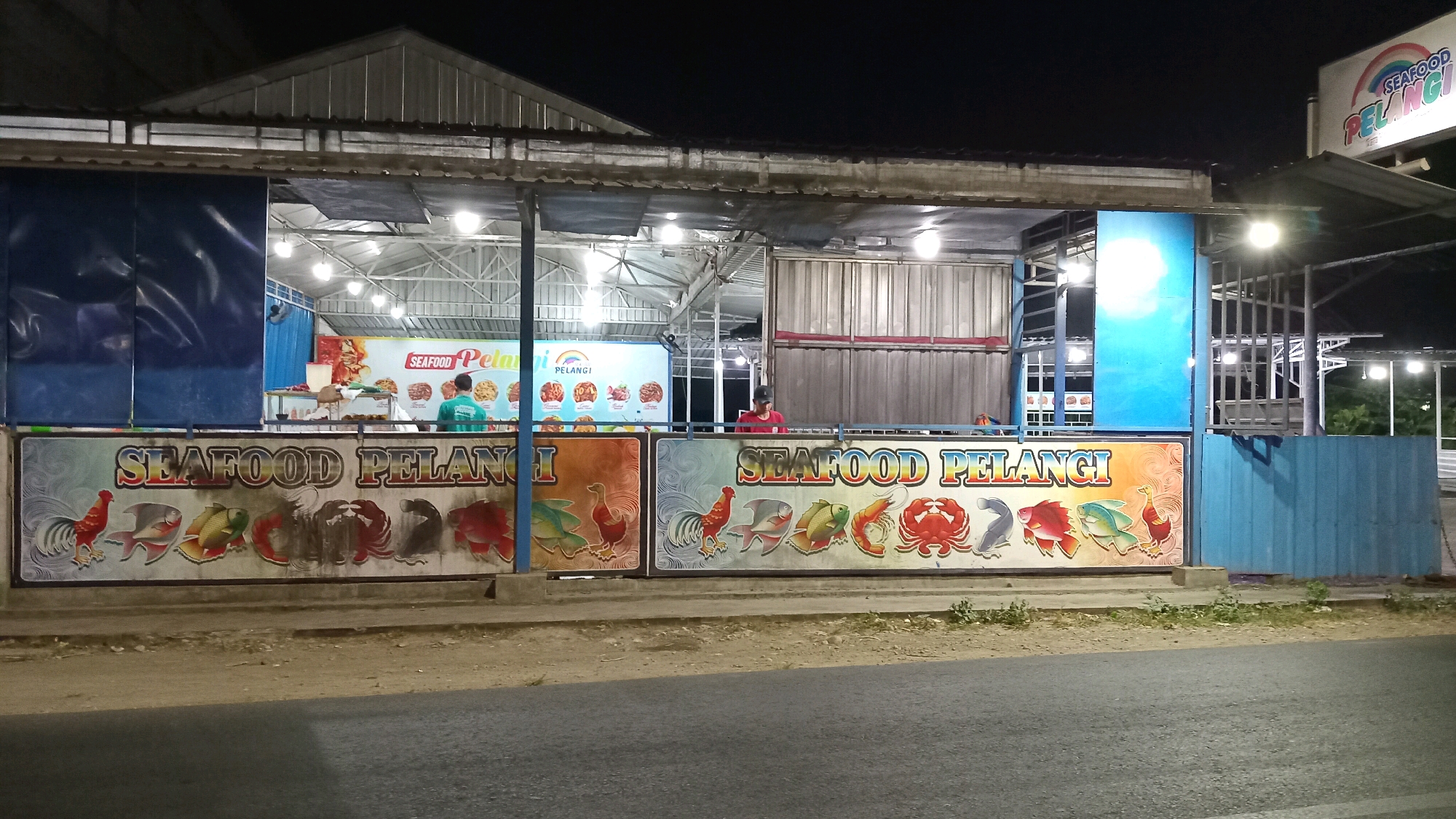 The image depicts a seafood restaurant named "SEAFOOD PELANGI" at night in Suci, Indonesia. The restaurant is an open-air structure with a corrugated metal roof supported by metal poles. The front of the restaurant features a large banner with "SEAFOOD PELANGI" written in large, colorful letters. Below this banner, there are two separate panels, each displaying various seafood illustrations, including fish, crabs, and a rooster.
Inside the restaurant, lights illuminate the area, revealing metal structures and what appear to be serving counters. Two individuals are visible: one wearing a green shirt and the other in a red shirt. The person in the green shirt is facing away from the camera and seems to be interacting with a customer or preparing food. The person in the red shirt is behind a counter, also facing away.
The restaurant is situated on a paved surface, possibly a road or parking area, with a sidewalk in front of the banner. To the right of the main structure, there's a blue metal wall and a sign that also reads "SEAFOOD PELANGI" with a rainbow graphic. Trees and foliage are visible in the background, suggesting a semi-urban or suburban environment. The overall impression is of a modest, roadside seafood eatery operating in the evening.