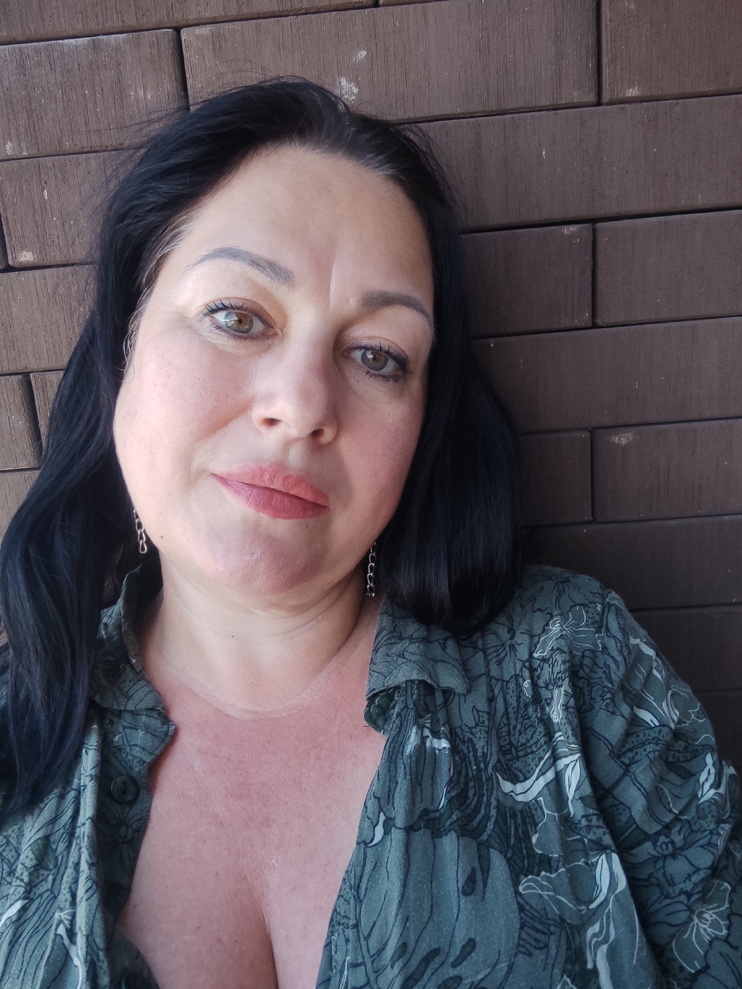 The media file is a medium close-up, selfie-style photograph of a woman with dark hair and brown eyes, leaning against a brick wall. She is wearing a green, floral-patterned shirt that is open at the collar, revealing her chest. The woman has on light makeup, including lipstick and eyeliner. She is wearing a silver earring in her visible ear. The brick wall behind her is dark brown and appears to be outdoors. The lighting suggests it is daytime, possibly late afternoon or early evening, with a warm, natural light. The woman's expression is neutral, with a slight smile. The overall mood is casual and relaxed. No text is visible in the image.