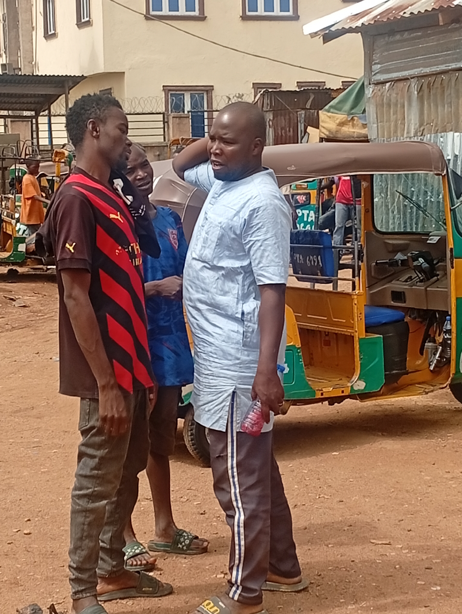 Three men are conversing in an outdoor setting in Jos, Nigeria. The man on the left is wearing a brown shirt with red and black stripes, jeans, and sandals. The man in the middle is wearing a blue soccer jersey, khaki shorts, and sandals. The man on the right is wearing a light blue shirt, grey pants with white stripes, and brown sandals. He is holding a plastic water bottle. Behind them is a yellow and green tricycle. The ground is dirt. In the background, there are buildings and other tricycles.