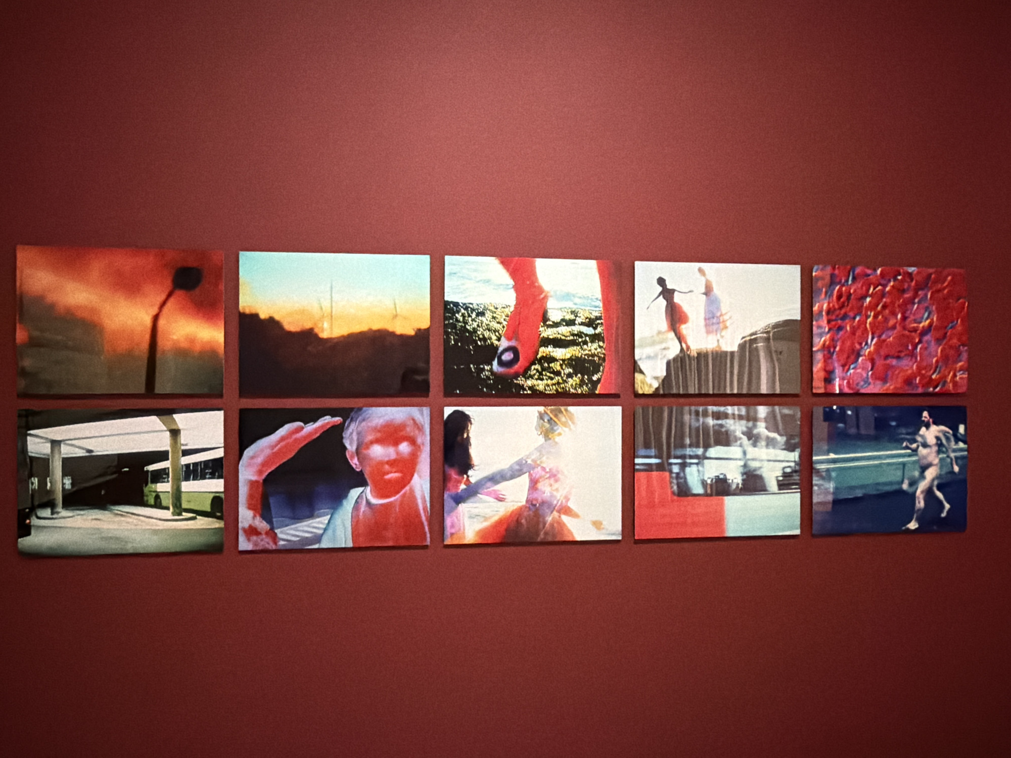The image shows a gallery wall with nine framed photographs arranged in a grid of three rows and three columns. The wall is painted a deep red.
The top row features three photographs. The leftmost photograph depicts a stylized image of a streetlamp silhouetted against a fiery orange and red sky. The middle photograph shows a dark landscape with silhouetted structures against a pale blue and yellow sky, with bright vertical elements suggesting distant towers. The rightmost photograph is an abstract close-up of bright red shapes, possibly flowers or fabric.
The middle row also contains three photographs. The leftmost image is a close-up of a bare foot with a red sock or stocking and a black toenail against a blurred background of what appears to be vegetation. The middle photograph shows two silhouetted figures of women in flowing dresses, one slightly in front of the other, on what looks like a raised platform or structure with a blurred background. The rightmost photograph is an abstract composition with a predominantly red foreground and a blurred background with hints of blue and grey.
The bottom row displays three more photographs. The leftmost image shows a bus shelter with a green bus visible in the background, illuminated by artificial light. The middle photograph is a close-up portrait of a child with an ethereal glow, the skin tones rendered in intense red and blue, and a hand raised near the face. The rightmost photograph shows two figures, one in the foreground with dark hair and wearing a brightly colored dress that appears to be in motion, and another figure behind it.
The bottom row's rightmost photograph depicts a man running towards the right side of the frame. He is unclothed and appears to be in motion on a dark, possibly wet surface, with a teal-colored barrier or wall to his left. The background is blurred, suggesting an urban environment with some indistinct structures.
The exhibition appears to be indoors, with the red wall being the primary background. No specific location within Mexico City is indicated by the content of the photographs.