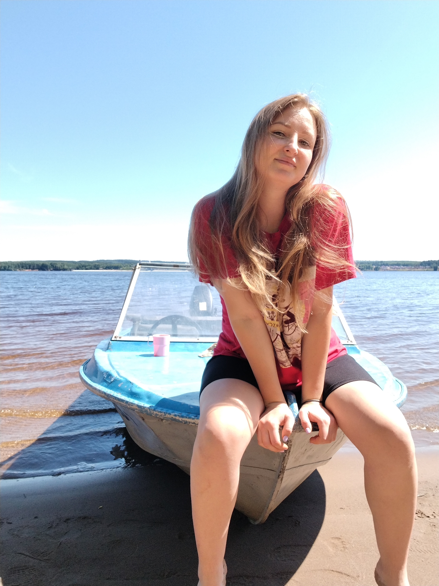 A young woman with long blonde hair is sitting on the edge of a small blue boat at the water's edge. She is wearing a red t-shirt with a graphic print and black shorts. The boat is pulled up onto a sandy beach, and the water appears calm with a distant tree-lined shore under a clear blue sky. The scene is set in Chaikovsky, Russia, on a sunny day.