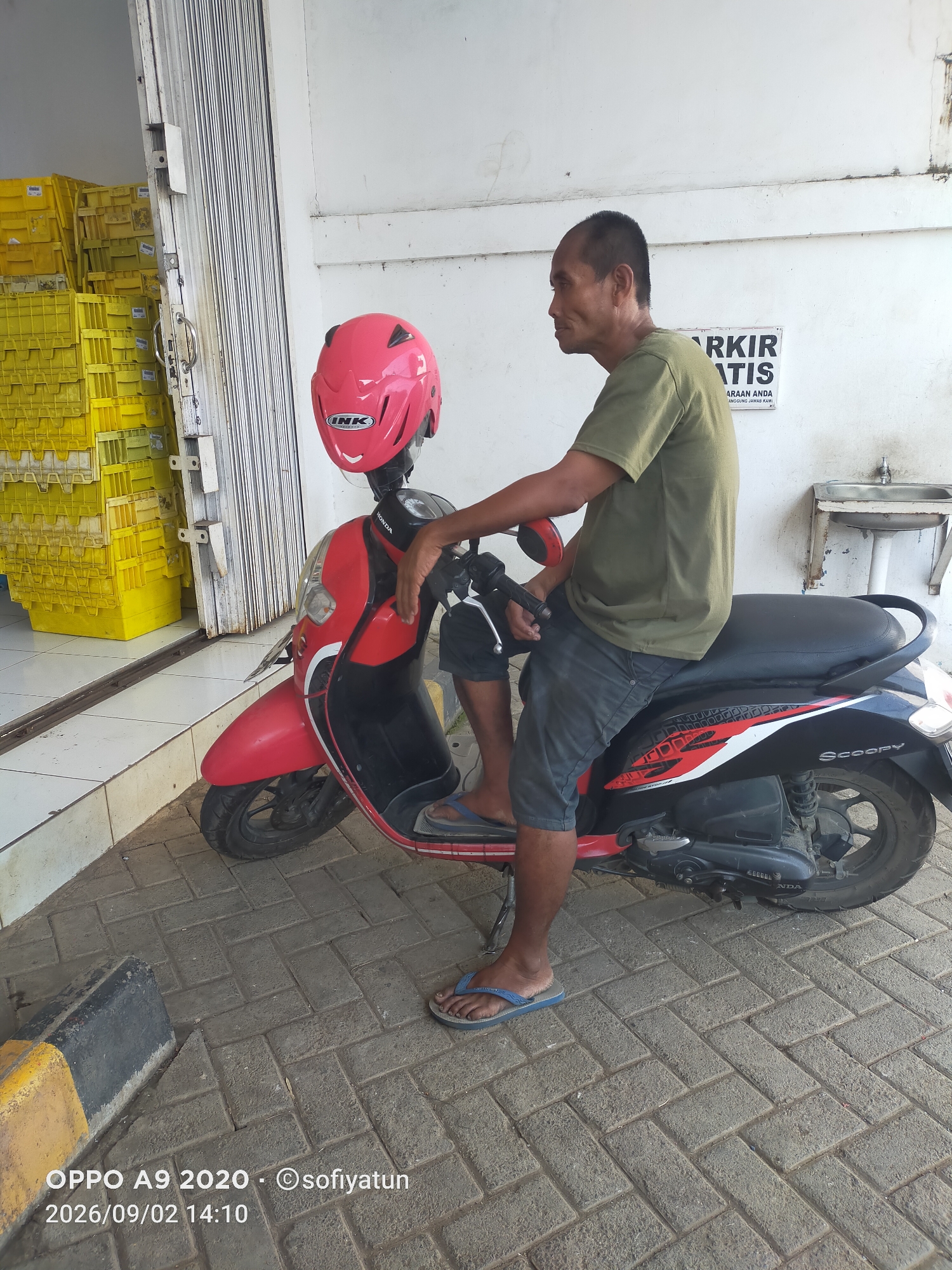A man sits on a red Honda Scoopy scooter. He is wearing a green t-shirt and grey shorts. A pink helmet rests on the seat behind him. The scooter is parked on a brick-paved area next to a closed metal roll-up door. To the left of the door are stacks of yellow plastic crates. A white wall with a sign that reads "PARKIR GRATIS" (Free Parking) is behind the man and the scooter. A metal sink is mounted on the wall to the right. The scene appears to be in an outdoor area of a commercial or storage facility. The timestamp on the image indicates it was taken on September 2, 2026, at 14:10. This location is identified as Tayu, Indonesia.