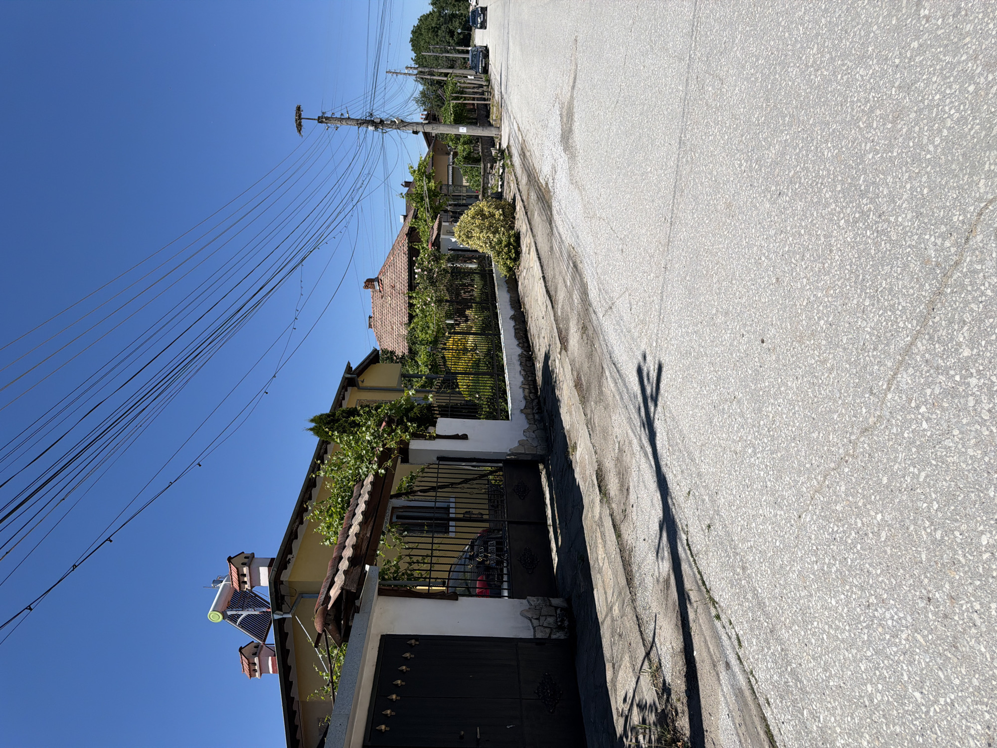 The image shows a street scene in Zlatitrap, Bulgaria, on a clear, sunny day. The street is paved with asphalt and has a sidewalk on the left side. The sidewalk is made of concrete and stone slabs. Along the sidewalk, there is a low stone wall with a black metal fence on top. Behind the fence, houses with red tile roofs are visible, with lush green vegetation and trees in their yards. Some houses have solar panels on their roofs. Telephone poles with a multitude of wires run along the street. A black car is parked on the right side of the street, and another dark-colored car is further down the road. The overall impression is of a quiet residential street in a rural or semi-rural setting.