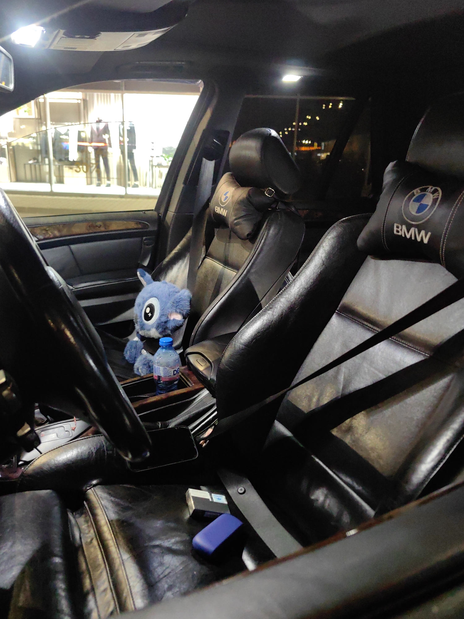 The interior of a BMW car is shown. The driver's seat and front passenger seat are both black leather with a BMW logo on the headrests. A blue stuffed toy, resembling the Disney character Stitch, sits in the driver's seat, next to a bottle of water. A blue object and a white box are on the driver's seat. The steering wheel is visible in the foreground. Outside the car window, a store with mannequins is visible and street lights suggest the scene is taken at night. The location is likely Sofia, Bulgaria.