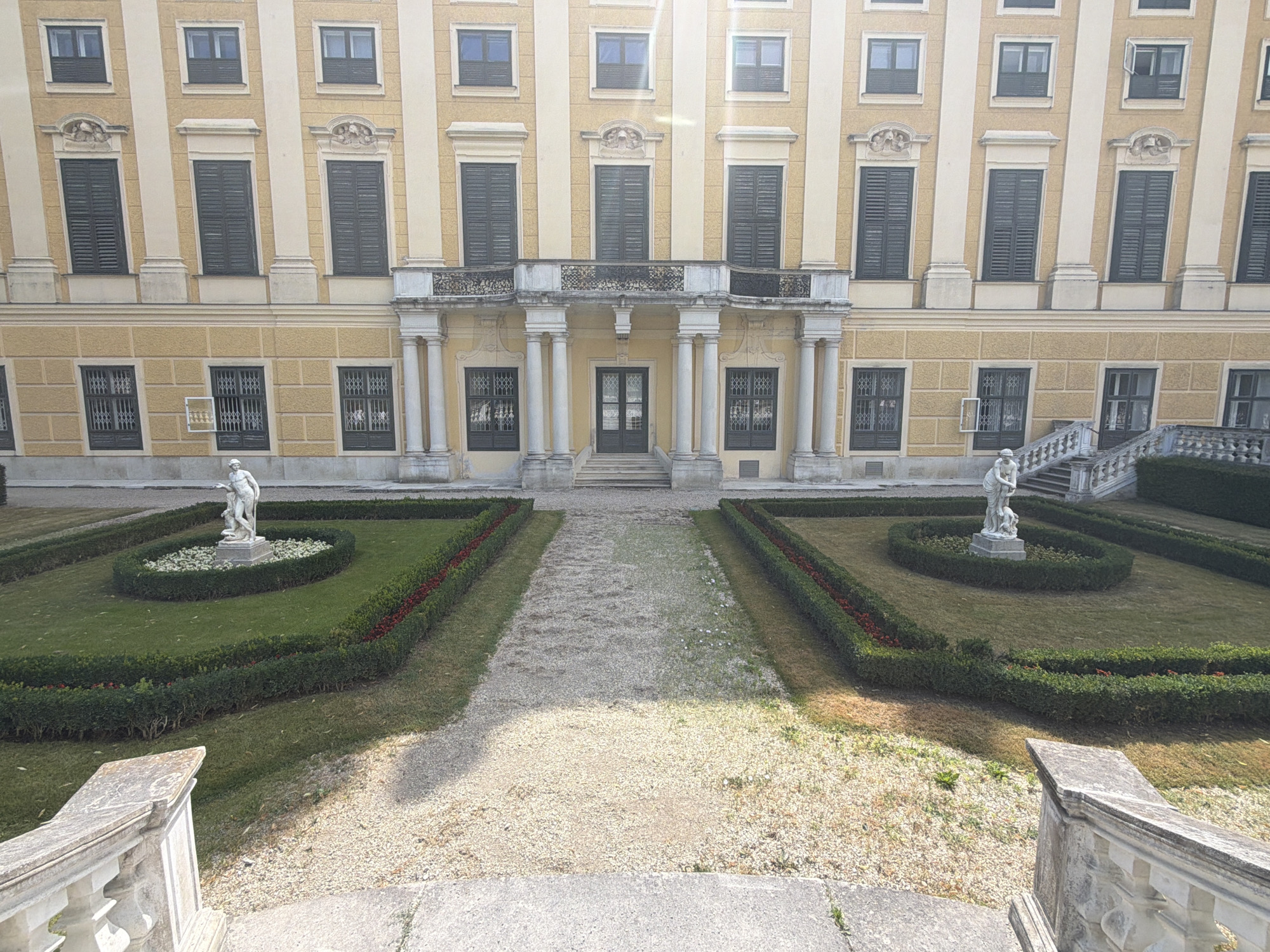 The image shows a view of Schönbrunn Palace in Vienna, Austria, looking towards its facade from a slightly elevated position. The palace is a large, yellow building with a neoclassical architectural style, featuring multiple rows of windows, some with dark shutters. A prominent central entrance is accessed by a set of stairs, flanked by columns and a balcony with an ornate railing.
In the foreground, a gravel path leads towards the palace, bordered by manicured green hedges with red flowers planted along their edges. Two oval-shaped garden beds are visible, each containing a white marble statue. The statue on the left depicts a seated female figure with a child, while the statue on the right is of a standing male figure. The gardens are well-maintained, with neatly cut grass. In the lower part of the image, stone balustrades of stairs and a pathway are visible, suggesting the viewpoint is from a similar elevated entrance. The sky is bright and clear, indicating a sunny day. There are no people visible in the scene, and no actions or interactions are taking place. The environment is one of a formal garden leading to a grand historical building.