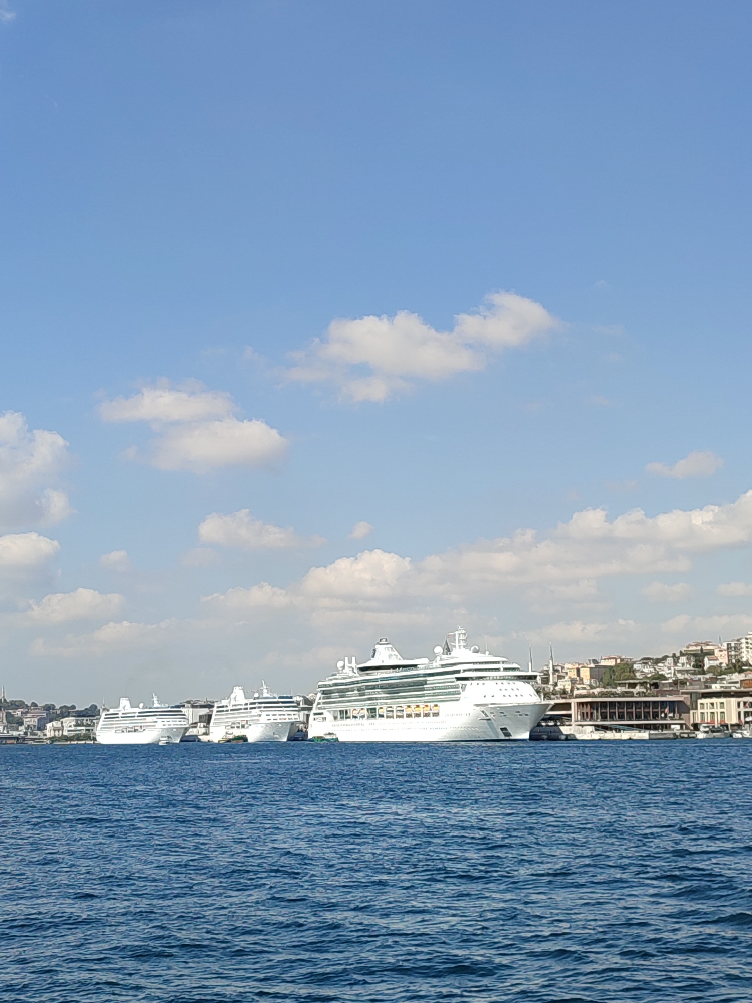 In the city of Beyoğlu, Turkey, a bright, sunny day is captured with a vast expanse of blue sky dotted with fluffy white clouds. The foreground is dominated by the deep blue, rippling waters of the sea. Along the coastline, a row of large, white cruise ships are docked. The largest ship on the right is prominent, with multiple decks and visible windows. To its left, two other similar cruise ships are lined up, appearing slightly smaller. In the background, a densely populated city landscape rises from the coast, characterized by buildings with terracotta roofs and a few taller structures. The water appears calm, reflecting the bright sky. No people are clearly discernible in the image.
