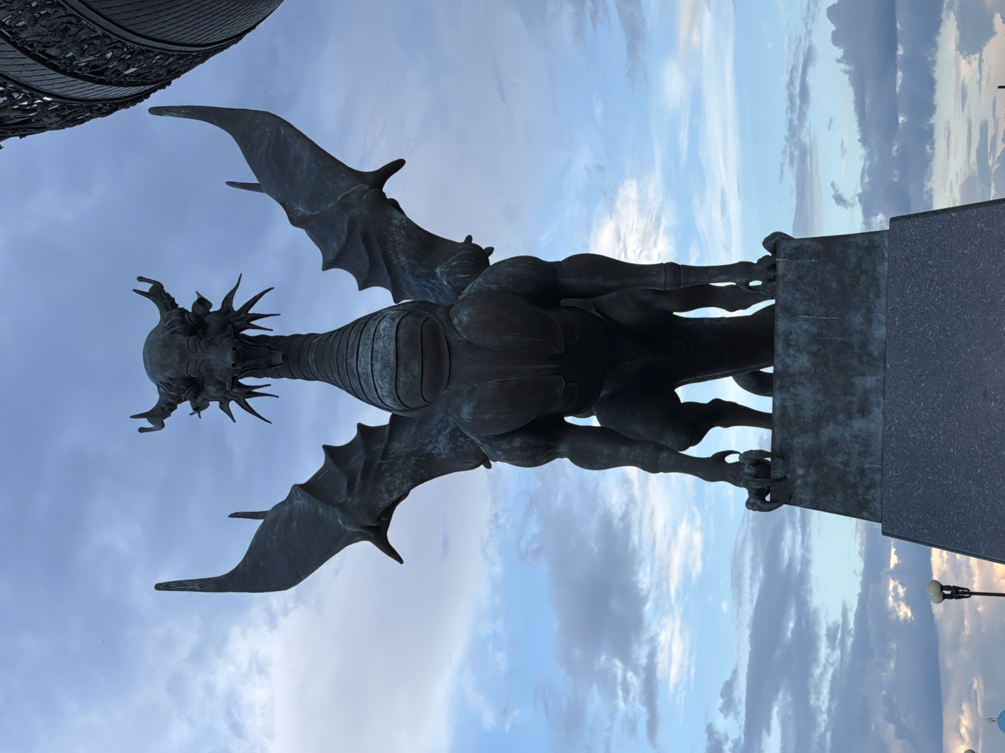 A dark, weathered bronze statue of a dragon is prominently featured against a pale blue sky with scattered white and grey clouds. The dragon, depicted with leathery wings outstretched and sharp claws, appears to be perched on a dark, rectangular stone pedestal. The statue has a detailed, somewhat menacing appearance, with a spiky crown around its head and a ribbed body. Its form suggests a powerful, mythical creature.
The perspective is from below, looking upwards at the dragon, emphasizing its imposing scale and its silhouette against the sky. The lighting suggests it is daytime, possibly late afternoon or early evening, given the soft light and the slightly muted colors of the sky. The overall atmosphere is one of grandeur and perhaps a touch of mystery, evoked by the dark, ancient-looking sculpture.
In the background, the upper portion of another large, dark, ornate metal structure is visible, possibly part of the same monument or a nearby building. Further in the distance, at the bottom of the frame, a streetlamp with a white light fixture is visible, and hints of other structures or lights can be seen, suggesting an urban setting. The clouds are varied, with some appearing wispy and others denser, indicating a dynamic sky. No text is visible in the image. The location is identified as Kazan, Russia.
