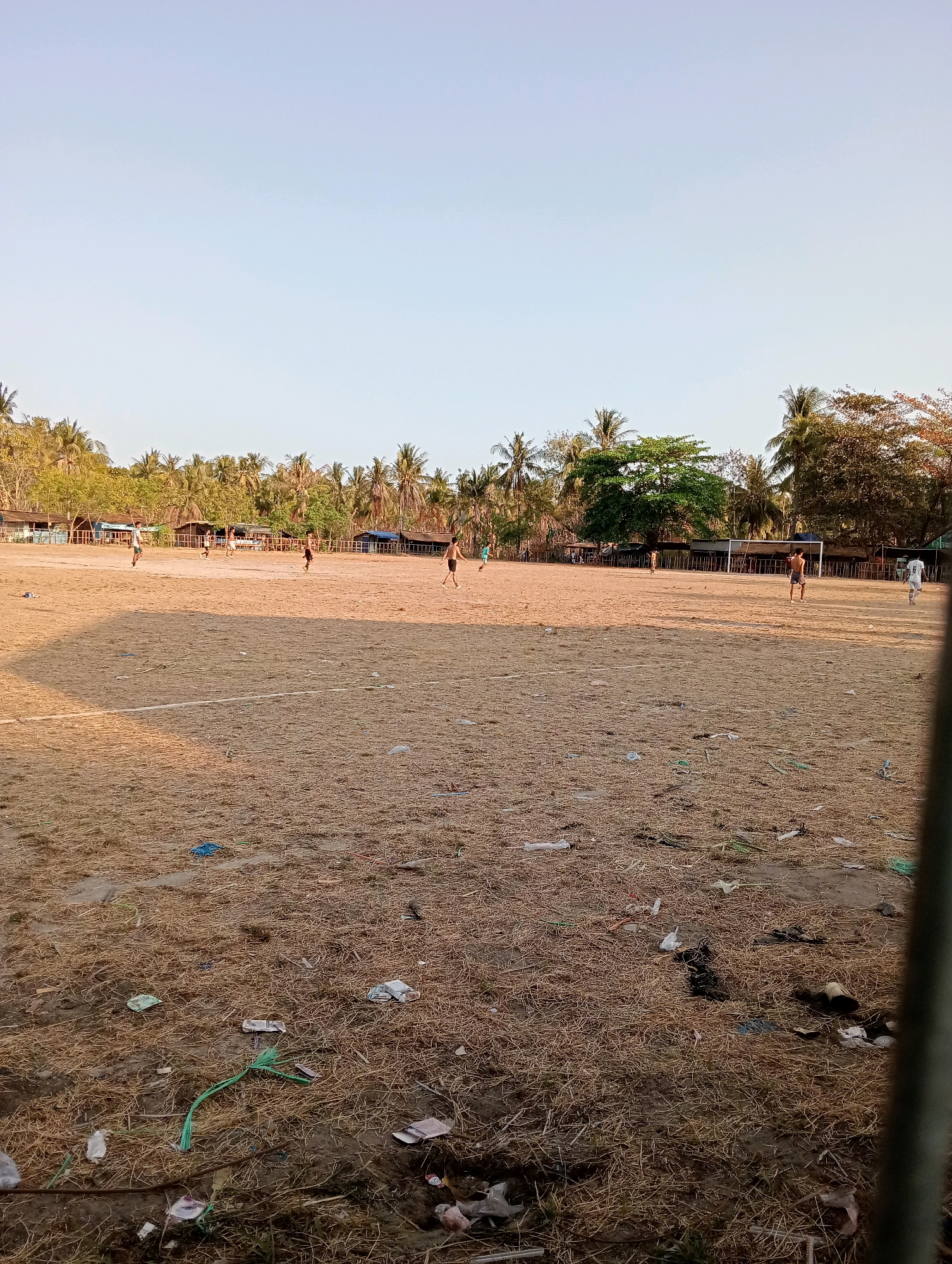 The image depicts a sparsely populated, sandy football field in Pagerungan Kecil, Indonesia. The field is dry and littered with debris, including plastic bags and dried vegetation. Several young men are visible on the field, appearing to be playing football. In the background, a line of small wooden structures, possibly houses or stalls, is visible, backed by a dense growth of palm trees and other vegetation. The sky is clear and light blue, suggesting it is daytime. The overall impression is of a rural or semi-rural setting.