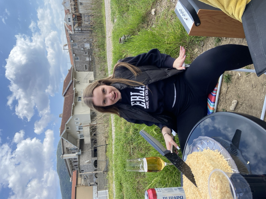 The image captures an outdoor scene in the city of Unknown, Unknown, featuring a young adult female seated at a table against a backdrop of residential buildings and mountains. The female subject, with long light-brown hair, wears a black "Everlast" sweatshirt, a black puffer vest, and black leggings. She is seated in a multi-colored striped folding chair, facing the camera with an open-mouthed expression and her hands gestured outwards. A glossy black circular table in front of her holds a round, light-colored cake with a crumb topping on a clear glass plate. A black-handled serrated knife is inserted into the cake. Also on the table are a tall champagne flute containing a yellow liquid, a red and white "Bavaria" branded beverage can, and a clear plastic cup with a dark liquid. To the subject's right, a wooden-sided speaker with silver controls is visible next to a dark-colored folding chair draped with a yellow fabric. The background consists of several multi-story houses with tiled roofs, some light-colored and others stone-clad, situated on a grassy, somewhat unkempt plot of land. Distant mountains rise under a partly cloudy blue sky.