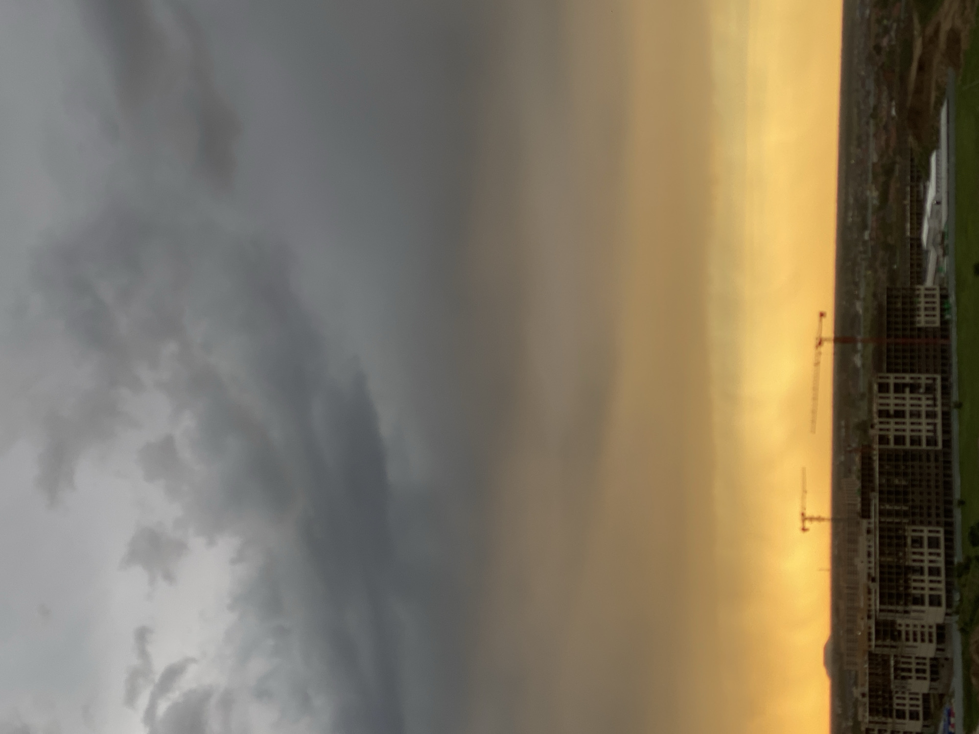 A wide-angle, full-shot photograph depicts a cityscape under a dramatic sky. The upper portion of the frame is dominated by dark, heavy clouds, tinged with gray and black, suggesting an impending storm or the aftermath of one. Below the dark clouds, the sky transitions to a band of warm, golden-orange light, likely from the setting sun, which illuminates the lower half of the image.
In the foreground, a construction site is visible. Several large, unfinished buildings, appearing to be apartment complexes or office buildings, are under construction. Their skeletal structures and exposed frameworks are evident. Red construction cranes are prominently positioned among these buildings, one with its boom extended horizontally, and another with its boom angled upwards. Green grass is visible in patches at the bottom of the frame, indicating a developed area or park. In the distance, a hazy horizon is punctuated by more low-rise buildings and a silhouetted mountain or hill on the left. The overall impression is one of urban development juxtaposed with the raw power of nature.