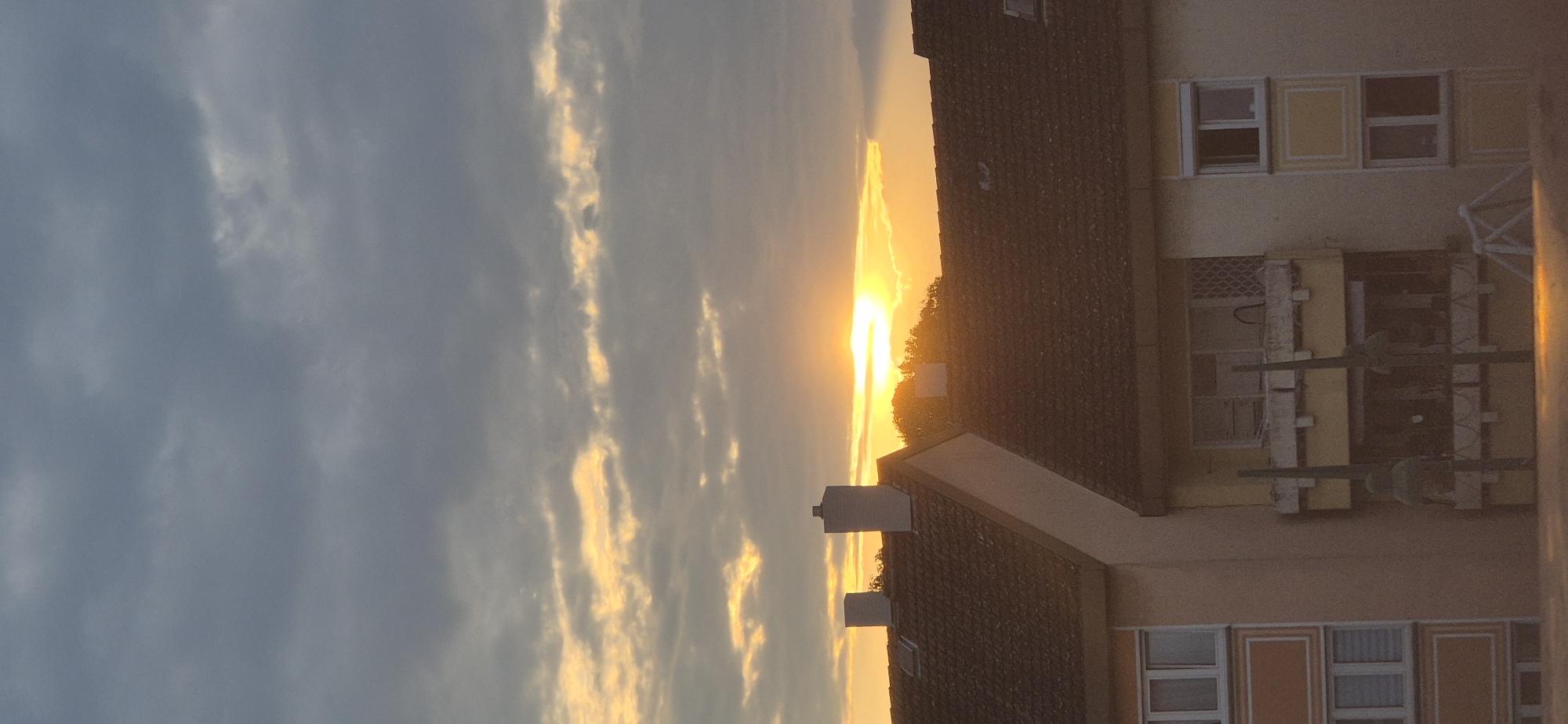 The image captures a sunset over the rooftops of a residential area in Mainz, Germany. The sky is filled with a mix of dark grey clouds and bright, golden streaks where the sun is setting, peeking through the clouds. Below the sky, the dark tiled roofs of buildings are visible. Two prominent chimneys stand on the roof in the foreground. Part of the facade of a yellow apartment building is visible on the right, featuring windows and a balcony. On the left, another apartment building with multiple floors and windows with orange trim is partially visible. The overall scene is of an urban landscape at dusk, with the warm light of the setting sun casting a glow over the rooftops and clouds.