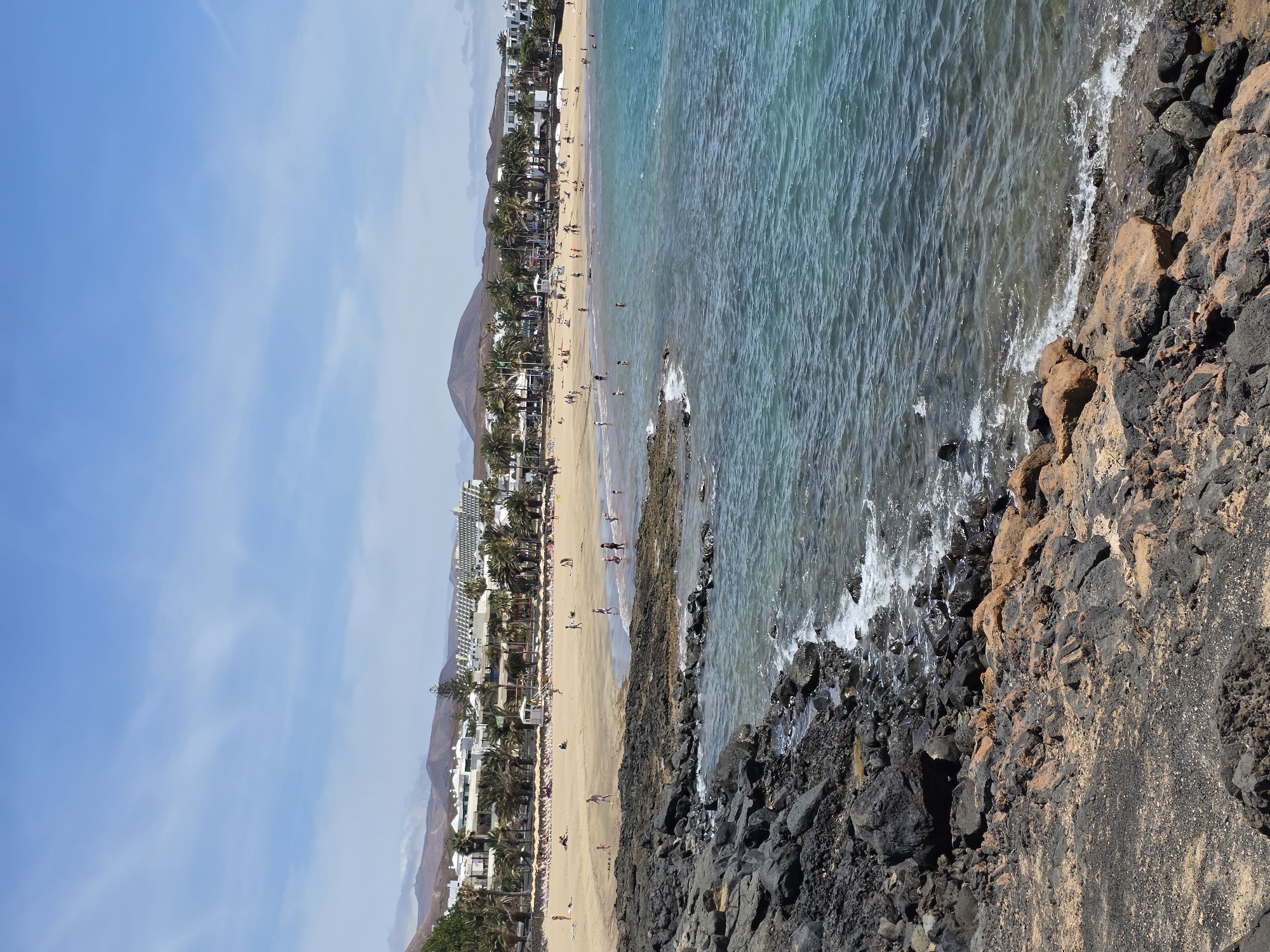 The image depicts a busy beach scene in Costa Teguise, Spain, under a clear blue sky with scattered clouds. In the foreground, dark volcanic rocks transition to sand. The beach is populated with numerous individuals sunbathing on loungers, walking along the shore, and wading in the turquoise ocean. A line of palm trees separates the beach from buildings in the background, which appear to be hotels or resorts. Mountains form a distant backdrop. The water is calm with gentle waves lapping against the shore.
