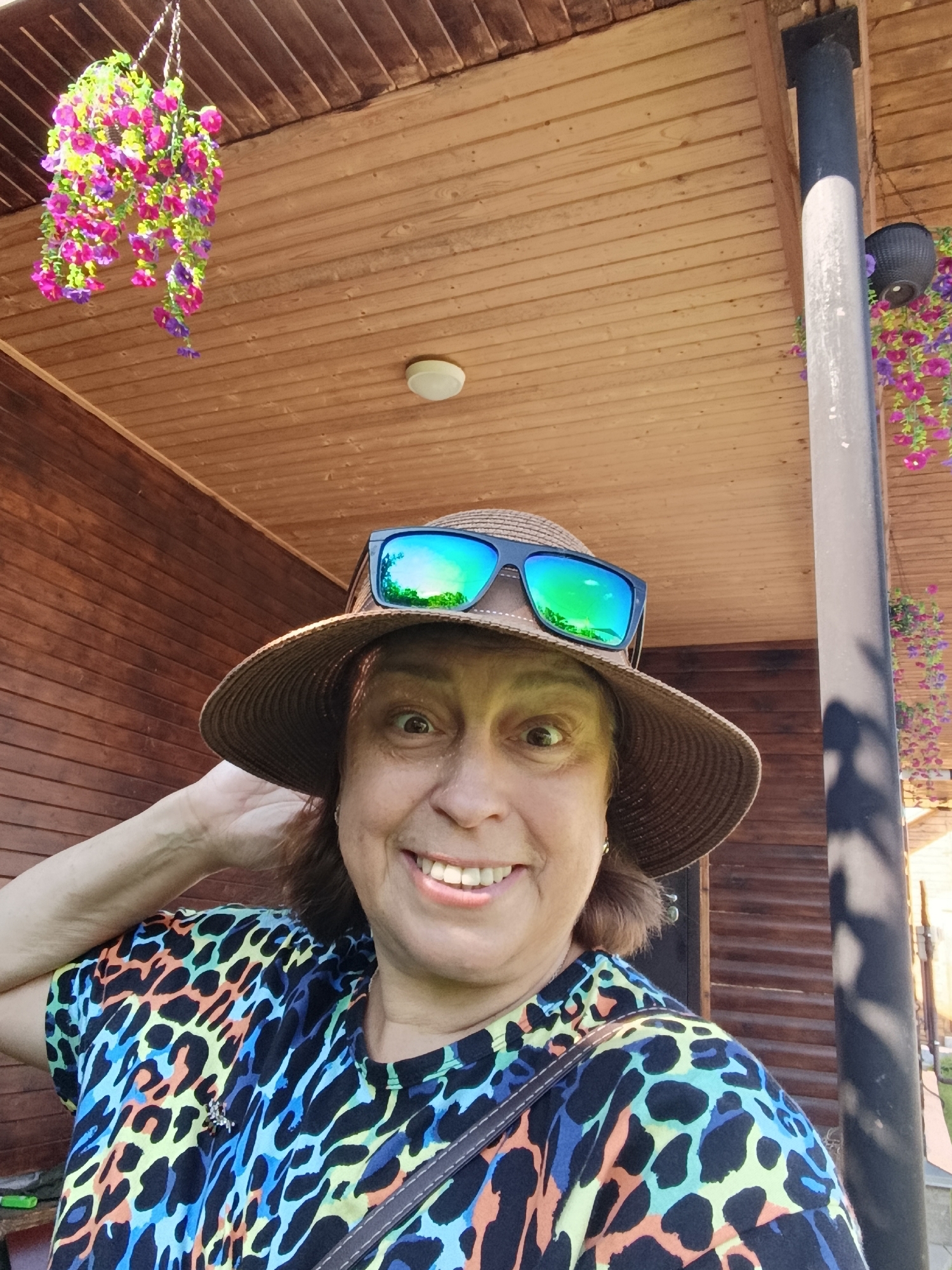 A medium-shot, selfie-style photo captures a woman with a wide smile, wearing a straw hat with sunglasses perched on top. Her eyes are wide open, looking directly at the camera. She has on a colorful, leopard-print t-shirt and is holding her left hand to her head. The background reveals a wooden porch ceiling with decorative hanging flowers. A dark-colored support pole stands to the right, casting a shadow on the wooden wall behind her. The scene suggests an outdoor setting, possibly a house or dacha in the city of Vereya, Russia.
