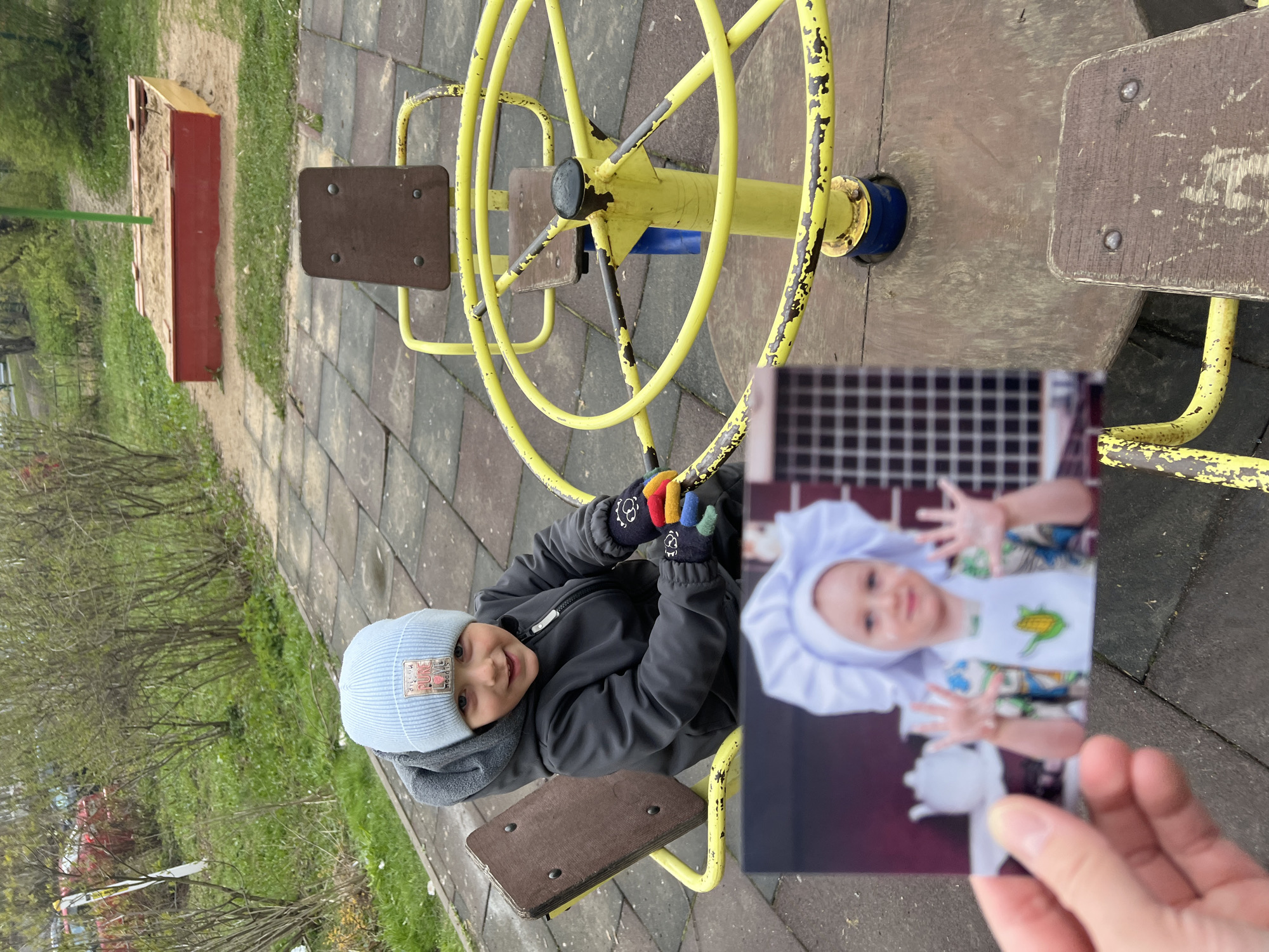 The image presents an outdoor playground scene in Mytishchi, Russia, characterized by overcast weather and bare trees, suggesting either early spring or late autumn.
The primary subject is a young child, likely a boy, sitting on a yellow metal merry-go-round. He is dressed warmly in a dark grey jacket, a light blue knitted beanie featuring a patch with the text "P.N.C." and "NEW," and multi-colored gloves (red, orange, green, and blue fingers with black palms adorned with white paw prints). He is looking directly at the camera with a cheerful smile. The merry-go-round itself shows signs of age, with yellow paint peeling to reveal a darker layer beneath.
In the lower right foreground, a human hand holds a printed photograph vertically. This photograph displays another young child, possibly a girl, dressed in a white ruffled chef's hat and a white apron with a green corn-on-the-cob design over a patterned shirt. Her hands are raised with open palms, and a small white teapot is partially visible at the bottom of the photo. The background of the photograph features a distinct dark grid pattern, suggesting it was taken indoors or in a studio setting.
The setting includes a paved ground made of dark square tiles. In the background, a red and yellow wooden sandbox is visible. Beyond the paved area and sandbox, there are patches of green grass and several leafless trees and bushes, indicating a park or residential outdoor space. The scene captures a moment of play on the playground, juxtaposed with a personal memory or different context presented in the held photograph.