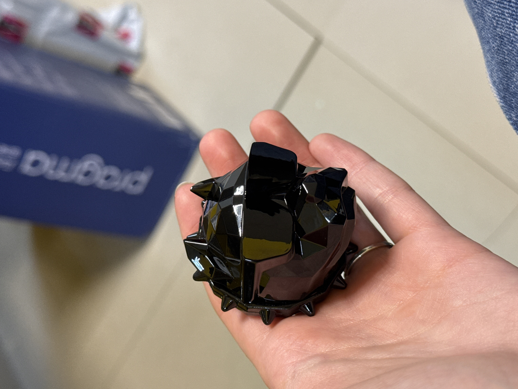 A black, geometric, spiky object is being held in a person's hand. The object is highly reflective. A blue box with white text that reads "DwGold" is out of focus in the background. The background also includes a light-colored tiled floor. The scene is indoors. The city and specific location are unknown.