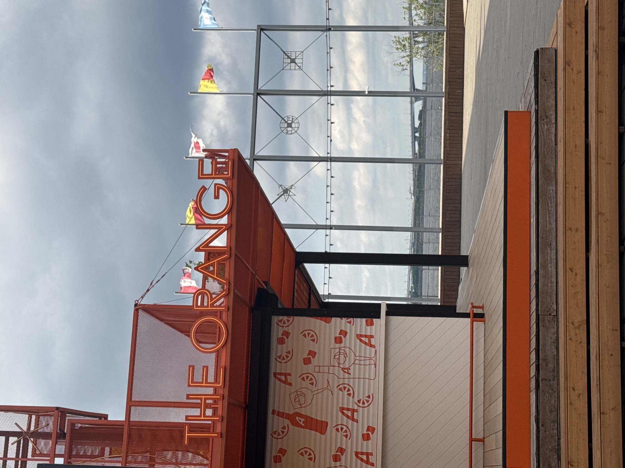 An outdoor scene in Saint Petersburg, Russia, features a red metal structure with a neon sign spelling "THE ORANGE". Below the sign, a white facade is decorated with red illustrations of bottles, wine glasses, and orange slices. Wooden decking surrounds the structure, with steps leading up to it. In the background, several flags are visible, along with a body of water and a bridge. The sky is overcast with dramatic clouds.