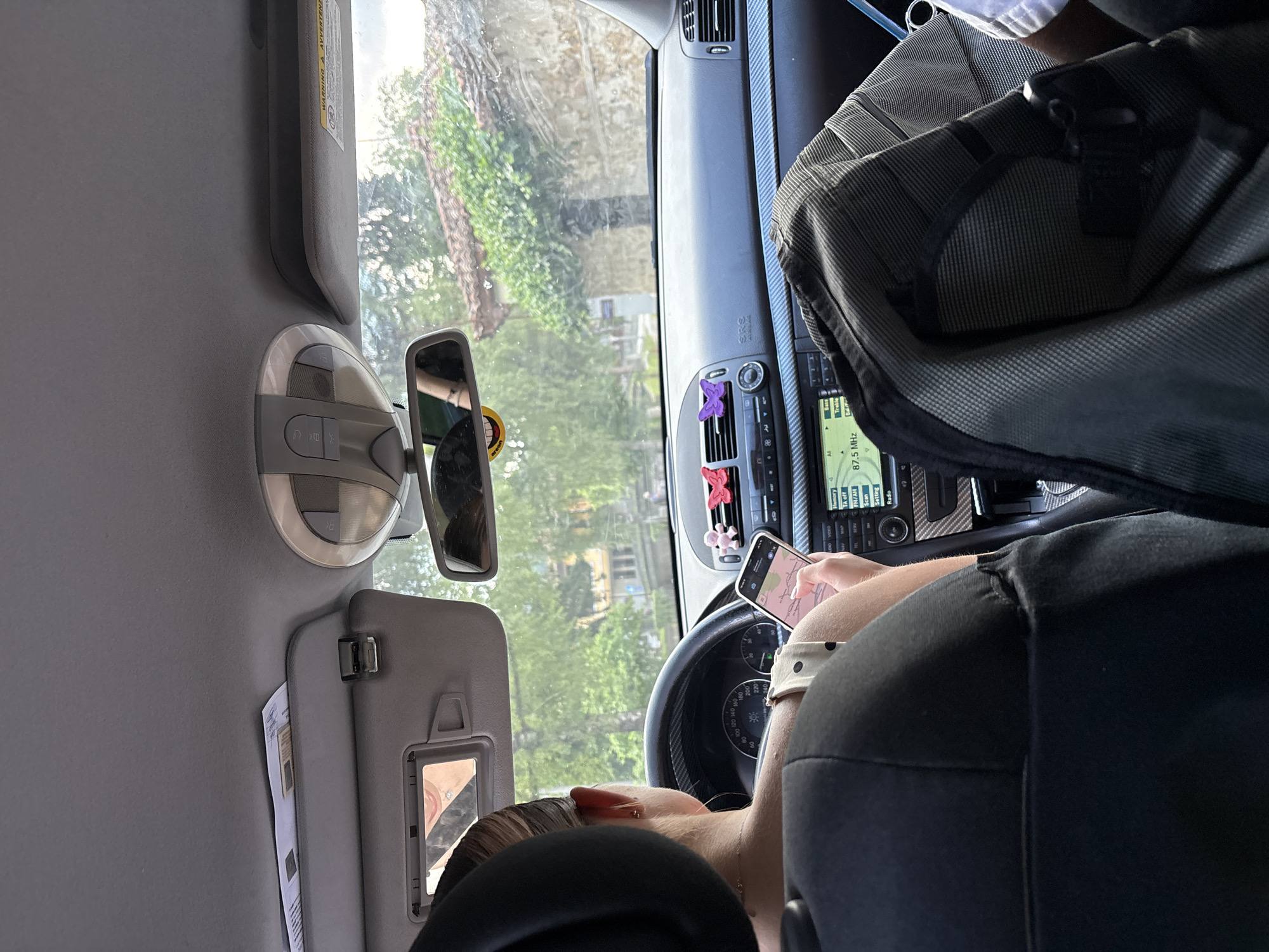 The image shows the interior of a car, with a person looking at their phone, which is displaying a map. The car appears to be in motion, as suggested by the blurred scenery outside the windshield and the car's dashboard. The view outside the window reveals trees and buildings, indicating a daytime setting, possibly a residential area or a scenic route. A backpack is visible on the passenger seat, partially obscuring the dashboard. The car's interior features a rearview mirror, a sun visor, and the car's audio system. The person's hand holding the phone, and the phone's screen with map details, are the main focus of the image. The visible text on the dashboard includes car information like "TA off", "FN/AN", "Setting", "Radio", and "87.5 MHz", along with a warning label in Italian ("AVVERTENZA"). The overall scene suggests a journey or a trip, with the driver or passenger using their phone for navigation. The lighting and clarity of the image suggest it was taken on a clear day.