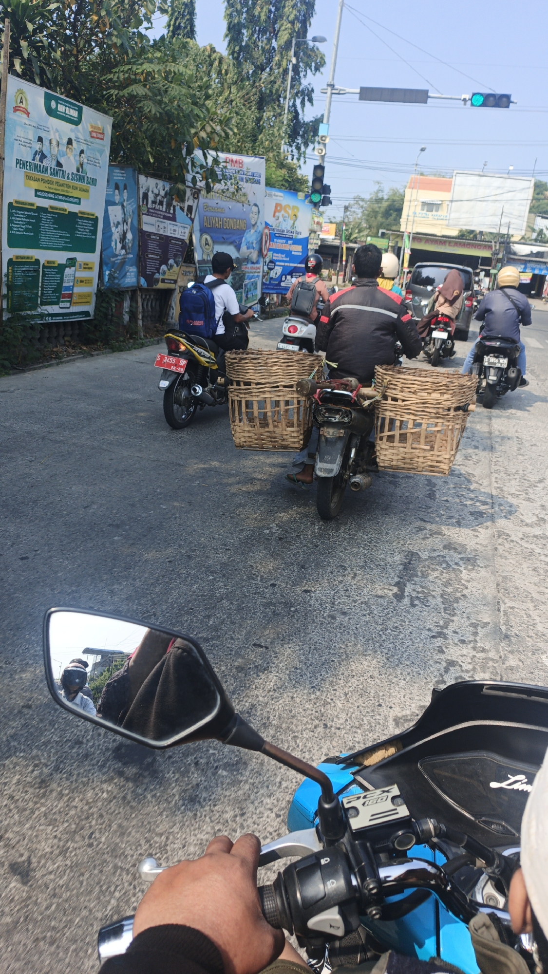 The image depicts a street scene in Pugeran, Indonesia, likely near a traffic intersection, as evidenced by the traffic lights visible in the background. The foreground shows a person's hand on the handlebar of a blue motorcycle, suggesting a first-person perspective. The motorcycle's side mirror reflects another motorcyclist wearing a helmet.

Further down the road, several other motorcycles are visible, some carrying passengers. Two motorcycles in particular are equipped with large woven baskets, one on each side of the rear. One motorcycle has a yellow frame and a license plate that reads "S 3453 SP 12-20". Another motorcycle, black in color, carries two prominent woven baskets. Riders on these motorcycles are wearing helmets.

In the background, there are various billboards, some with text and images that appear to be advertisements or public notices. One billboard has text in Indonesian, mentioning "PENERIMAAN SANTRI & SISWA BARU". Trees line the side of the road, and buildings can be seen in the distance. The sky is clear and blue, indicating a sunny day. The road surface is asphalt and appears to be in moderate condition. The overall scene suggests a typical day of transportation and commerce on Indonesian streets.
