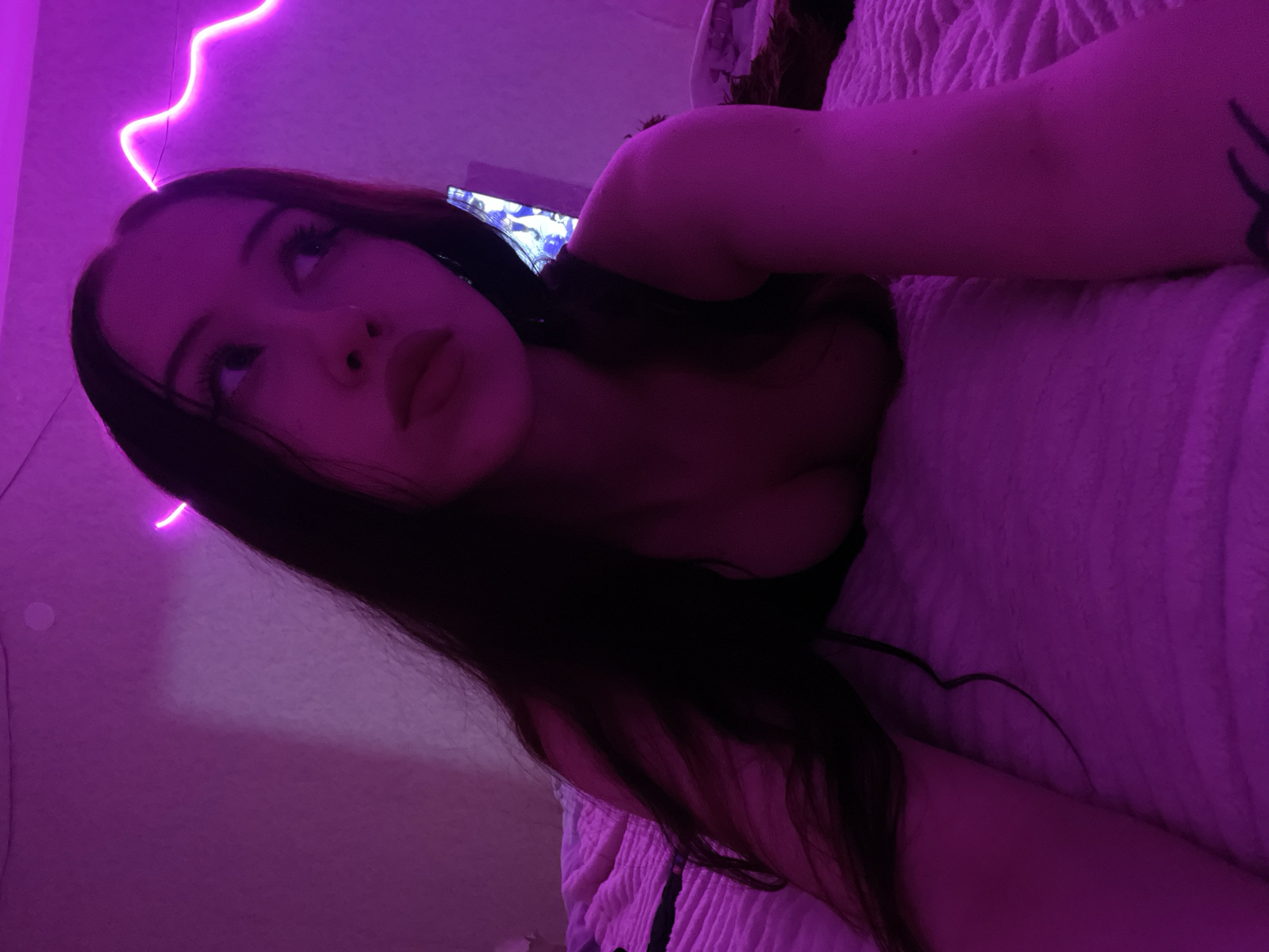A young woman with long, dark hair is lying on a bed in a room illuminated by purple neon lights. Her head is tilted and she is looking to the right, with her lips slightly parted. She is wearing a dark-colored top. The bed is covered with a textured pink blanket. In the background, a television screen is visible with a blue and white image. The overall scene suggests an indoor setting with a focus on mood lighting.