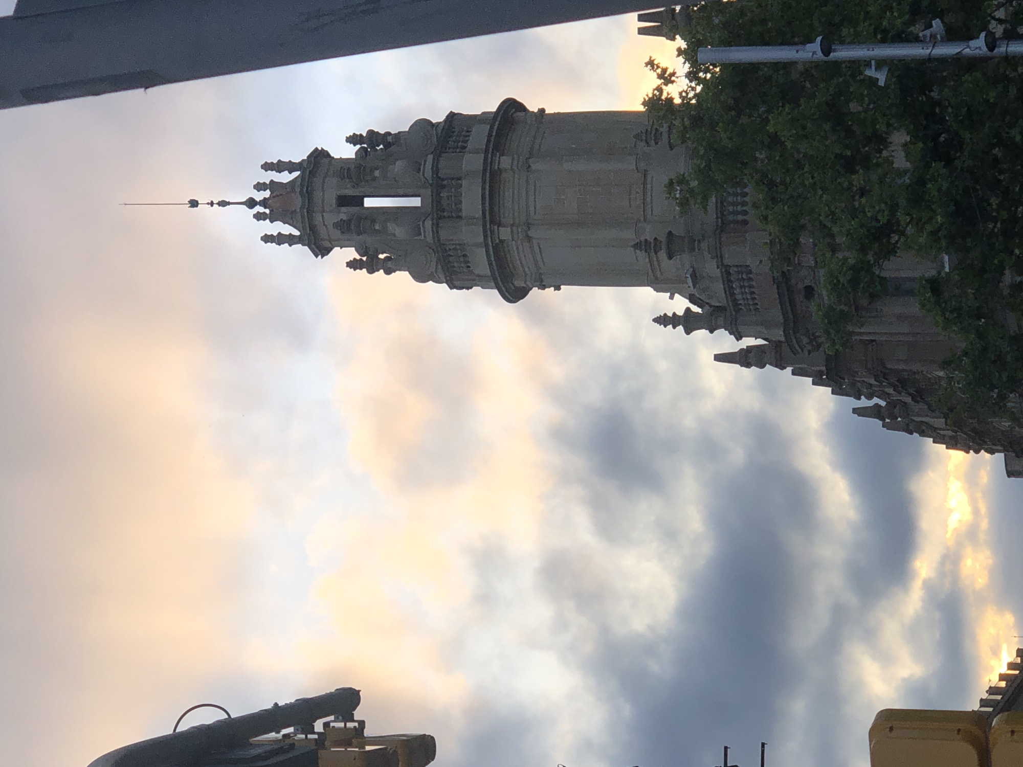 A tall, ornate stone tower with a pointed spire rises against a sky filled with dramatic clouds, tinted with hues of orange and yellow from the setting sun. The tower is detailed with decorative elements, including balconies and smaller spires. To the left, a portion of a yellow traffic signal and some overhead infrastructure are visible, suggesting an urban setting. Green foliage from trees partially obscures the lower section of the tower and the building it is part of. The overall scene evokes a sense of grandeur and the approaching end of the day in an urban landscape.