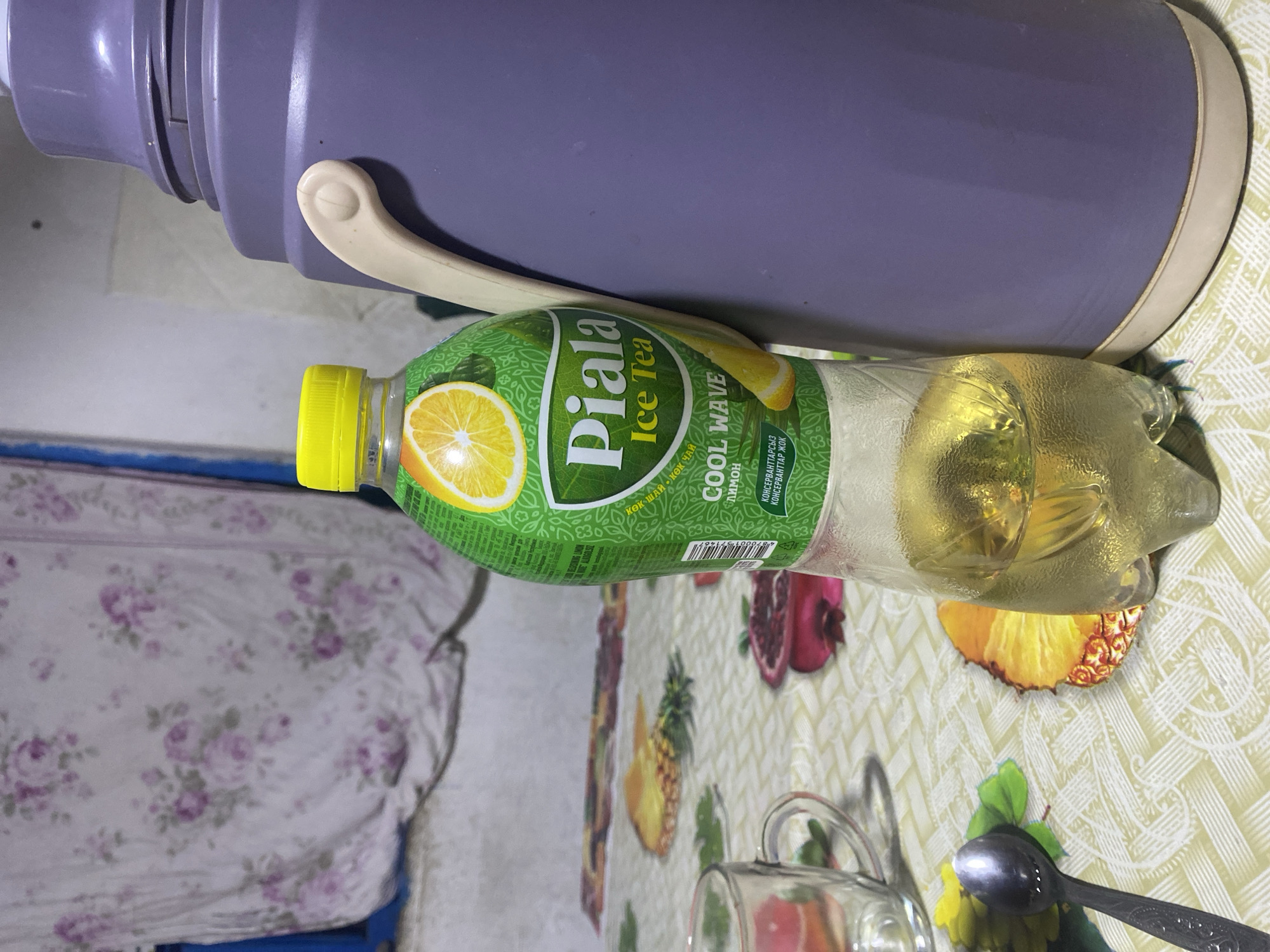 In Dostuk, Kyrgyzstan, an indoor scene depicts a table covered with a patterned tablecloth featuring images of fruit slices, including pineapple and pomegranate. On the table, a clear glass mug is visible, positioned to the left of a bottle of "Piala Ice Tea" with a lemon flavor. The bottle contains a light yellow liquid. To the right of the bottle stands a large, purple thermos with a beige handle. A silver spoon rests on the tablecloth to the left of the mug. In the background, a piece of fabric with a floral pattern hangs, possibly a curtain or bedsheet. The overall impression is that of a casual, domestic setting.