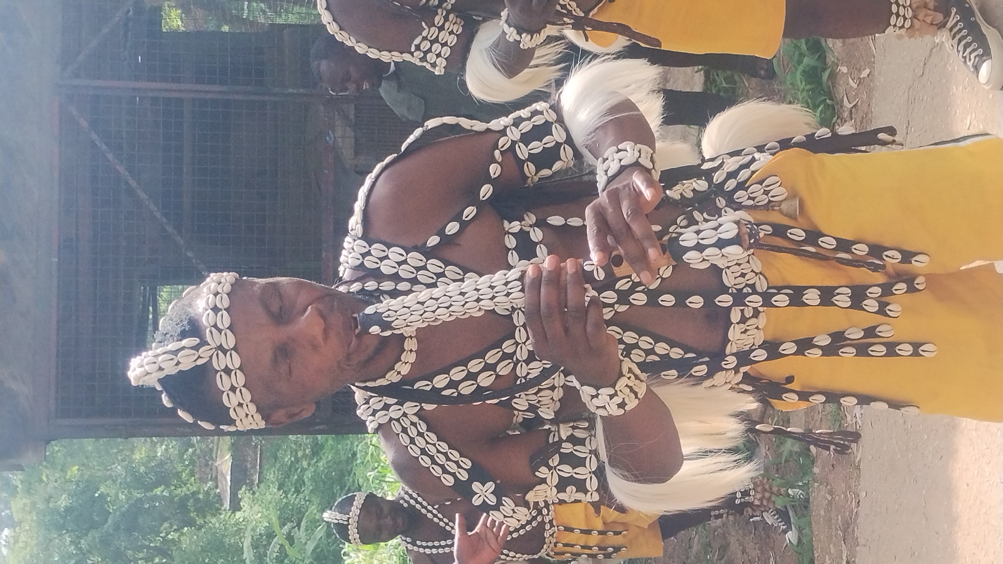 The image captures a group of people adorned in traditional attire, possibly for a cultural performance or ceremony. The primary subject is a man in the foreground, wearing a headdress and elaborate body ornamentation made of shells and dark fabric. He is holding a wooden instrument, possibly a flute, and appears to be playing it. His eyes are closed, suggesting concentration or immersion in the music.
The man's attire includes a yellow garment, possibly a wrapper or skirt, and a chest piece adorned with strings of shells. Similar shell decorations are visible on his wrists and head. Another individual, also in traditional dress, is partially visible in the background, their face turned away.
The setting appears to be outdoors, with hints of a rustic or natural environment. In the background, a metal fence structure and some greenery suggest a partially constructed or open-air venue, possibly in Jos, Nigeria. The lighting suggests it is daytime, with bright sunlight casting shadows. The overall mood is one of cultural engagement and performance. There is no visible text in the image.