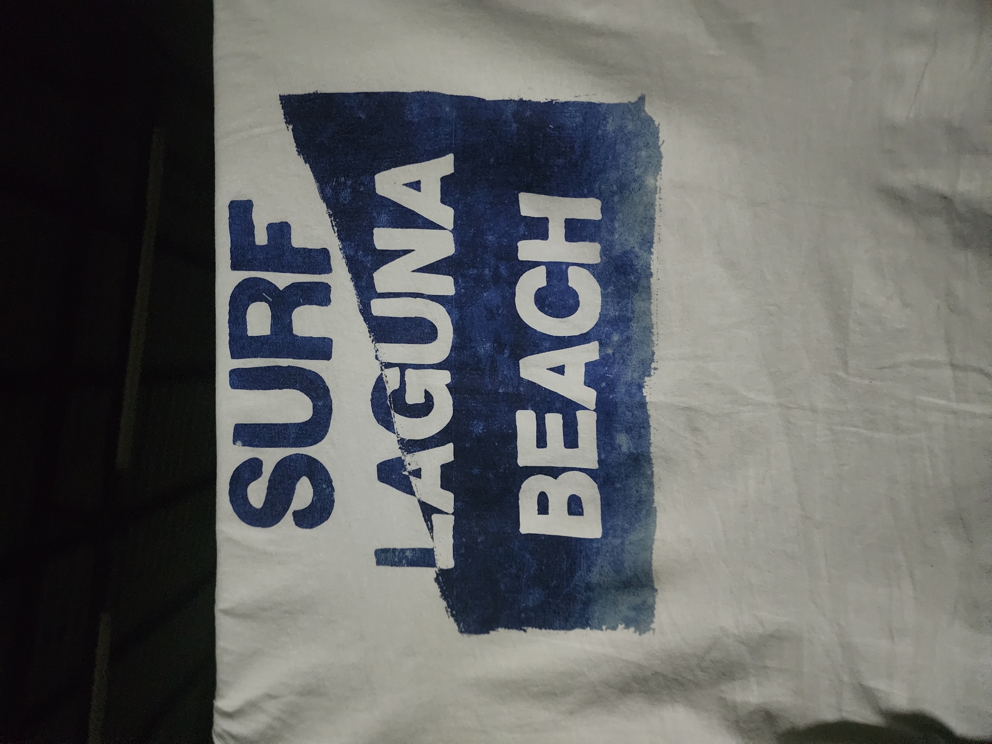 A white t-shirt is laid out flat. Printed on the t-shirt are the words "SURF LAGUNA BEACH" in bold, dark blue font. A rough, abstract blue shape, resembling a wave or a mountain range, is placed behind and slightly overlapping the words "LAGUNA" and "BEACH." The t-shirt is slightly wrinkled, and the lighting creates shadows on the fabric. The image does not depict any scenes or landmarks in Ushtobe, Kazakhstan.
